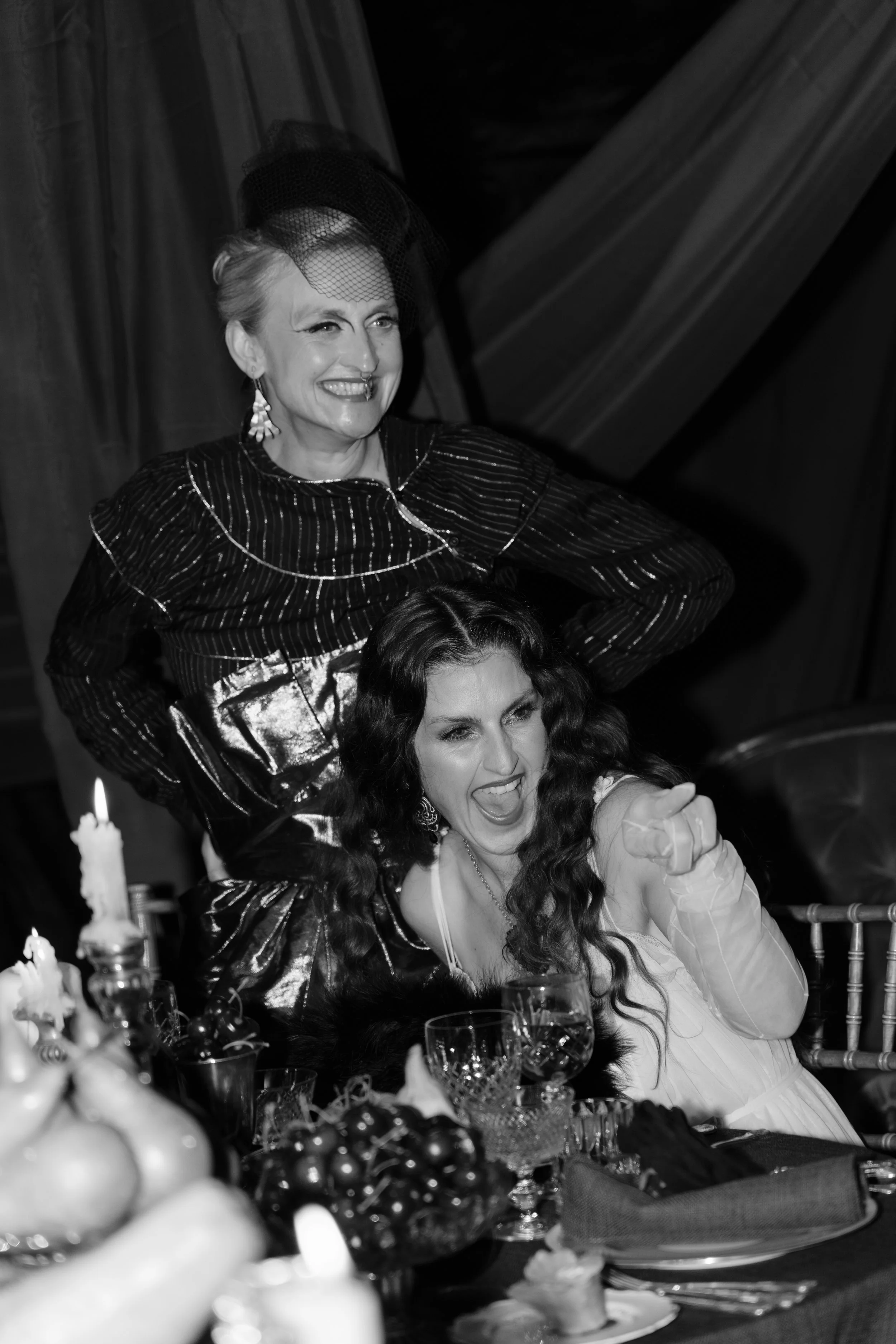 Two women are at a celebratory event, one standing and smiling while the other sits, pointing and yelling excitedly. The table in front has candles, grapes, and glasses.