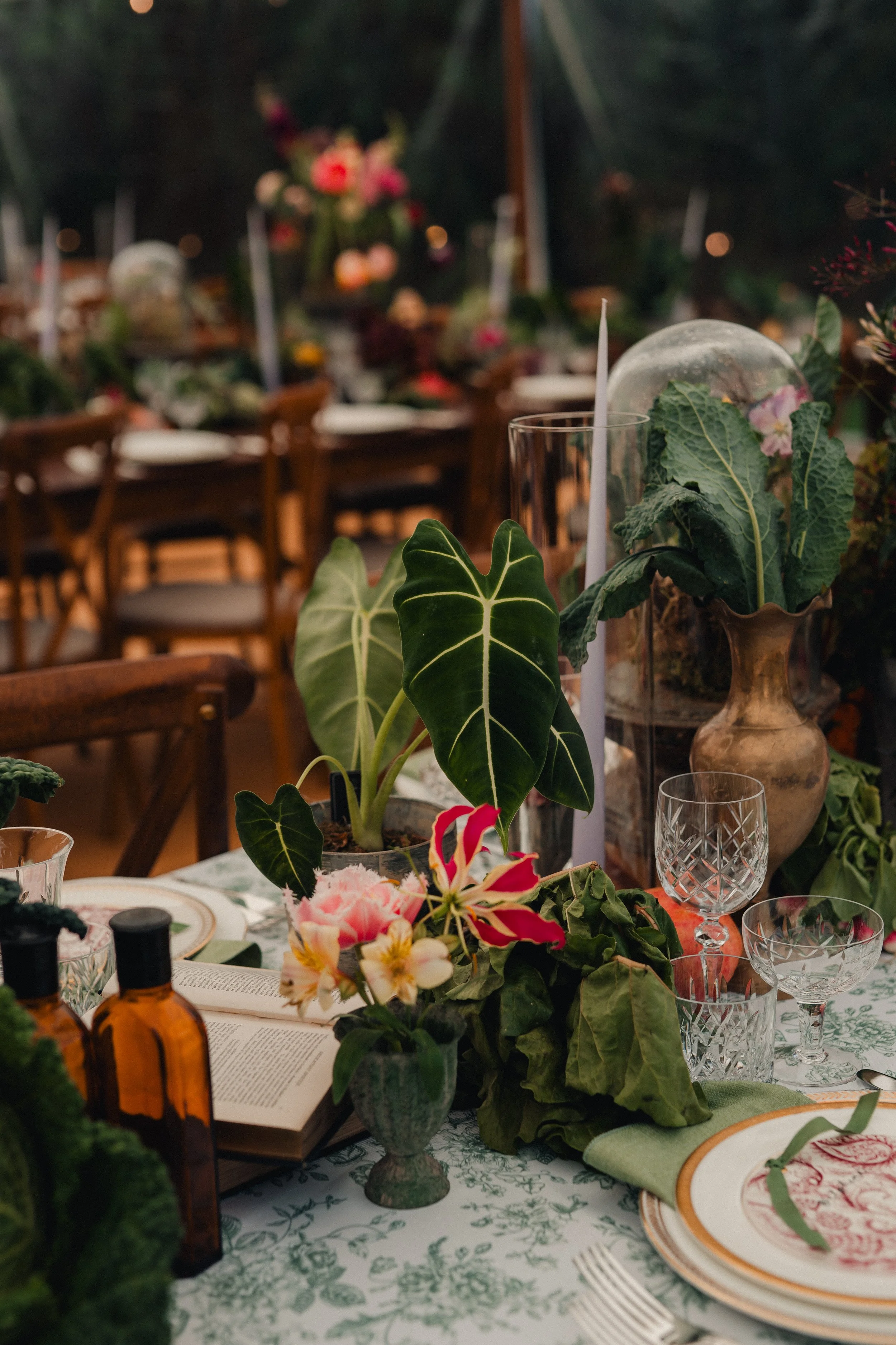 Decorative table setting with green plants and flowers, glassware, and plates, at an indoor event with a floral motif and wooden chairs in background.