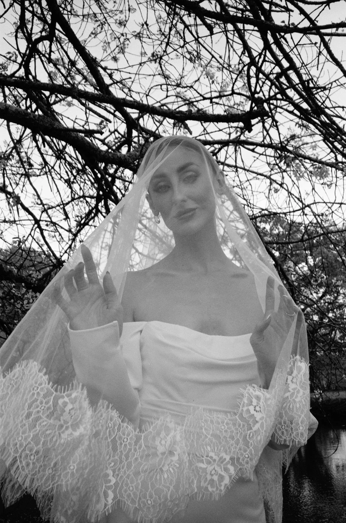 A woman in a wedding dress with a veil, standing outdoors under tree branches, with her hands raised near her face.