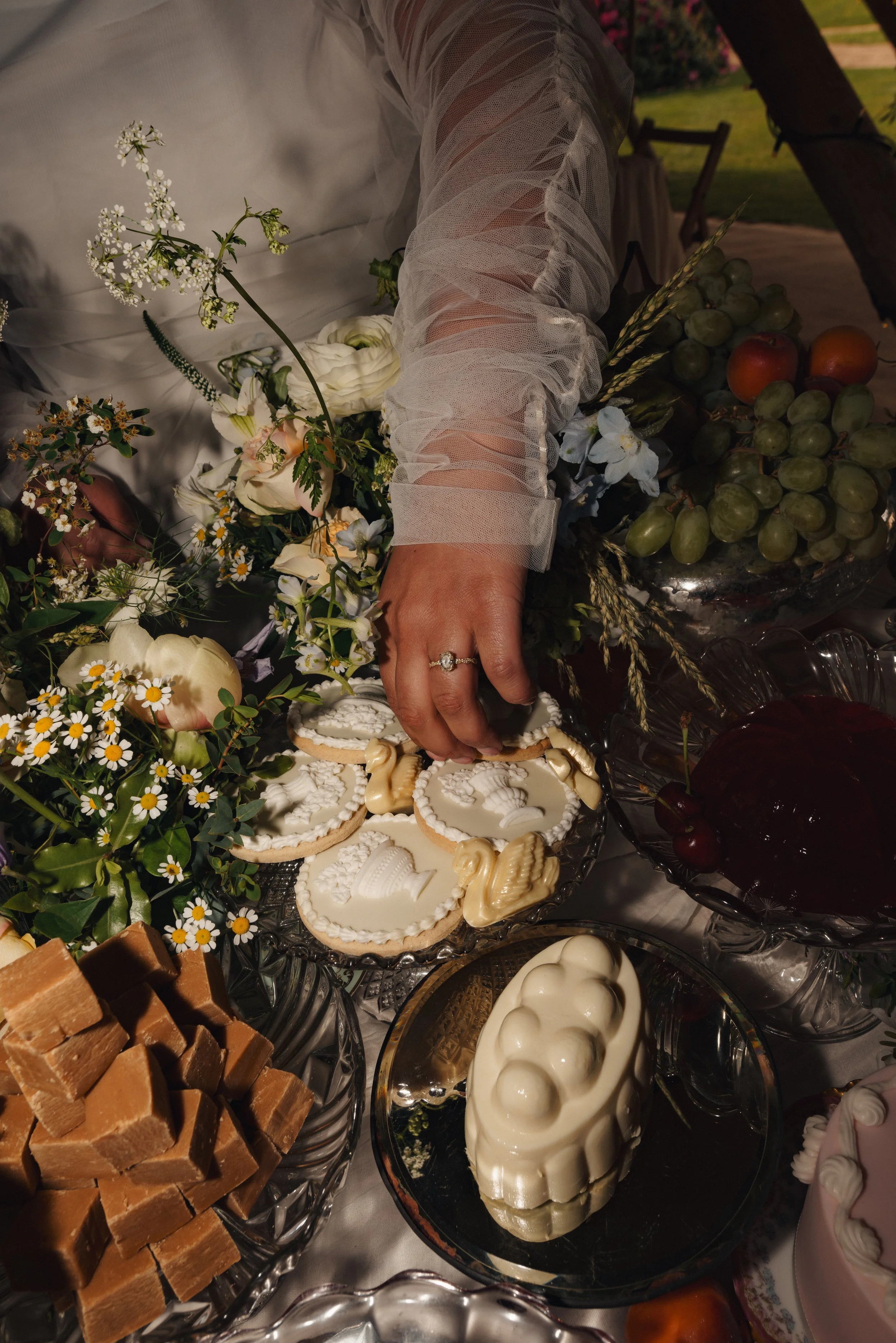 A person reaching for cookies on a dessert table filled with various sweets, flowers, and fruits during a celebration or event.