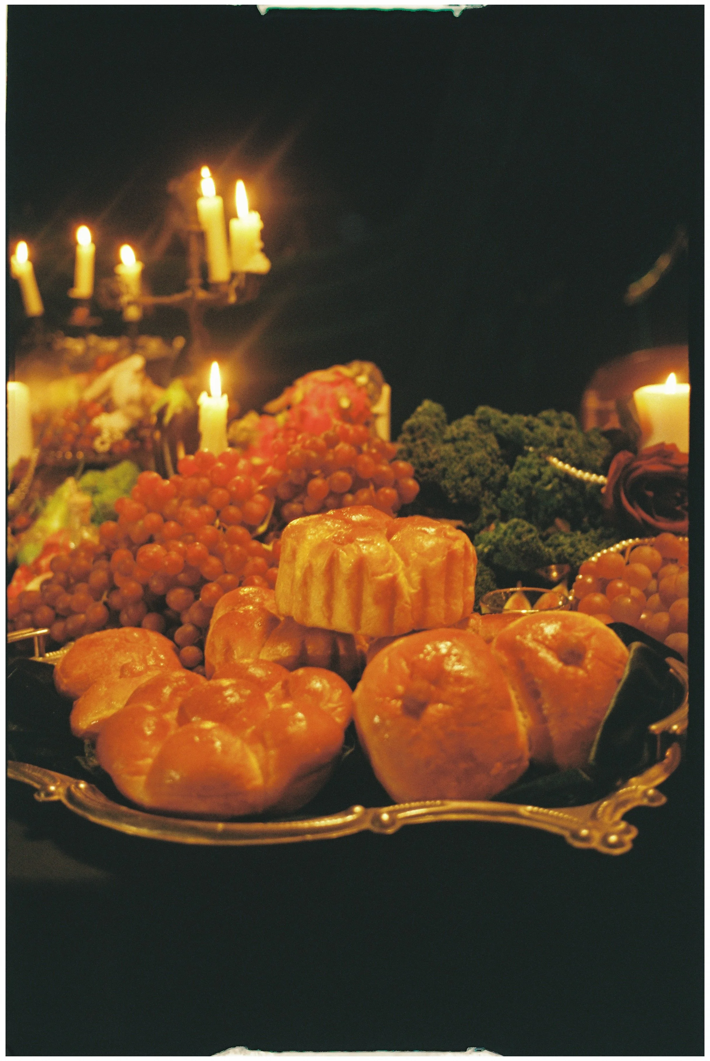 A silver tray with various baked goods, surrounded by grapes, broccoli, candles, and dark floral decorations.