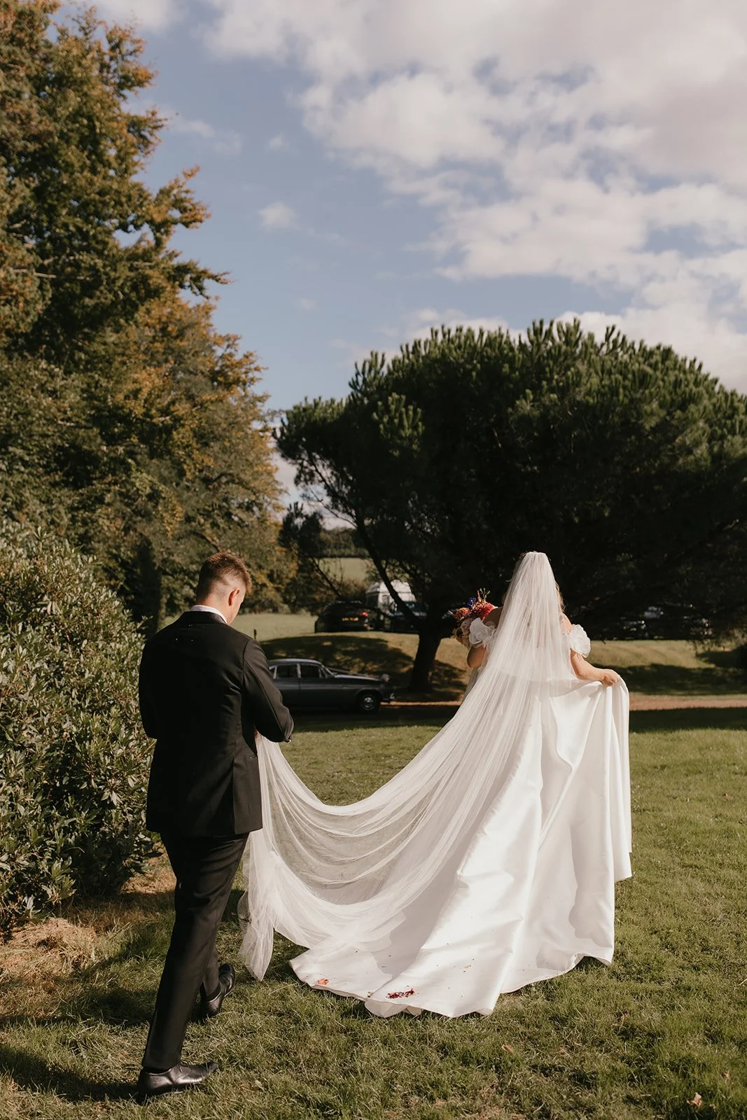 A bride in a white wedding gown and veil walking outdoors with a man in a black suit, holding her train, on a grassy area with trees and cloudy sky in the background.