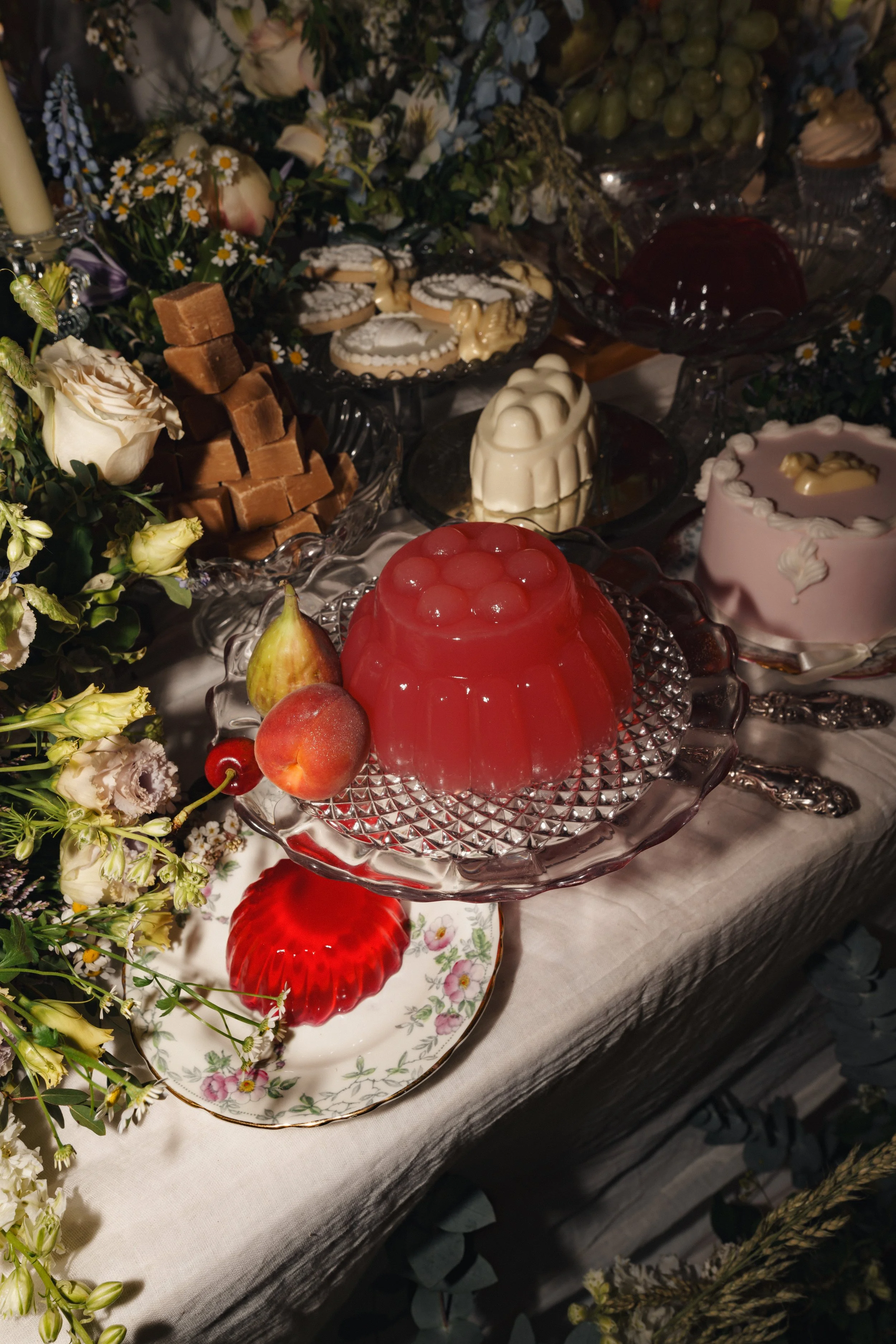 Assorted desserts including red jelly, biscuits, chocolate cubes, and confections on a table decorated with flowers.