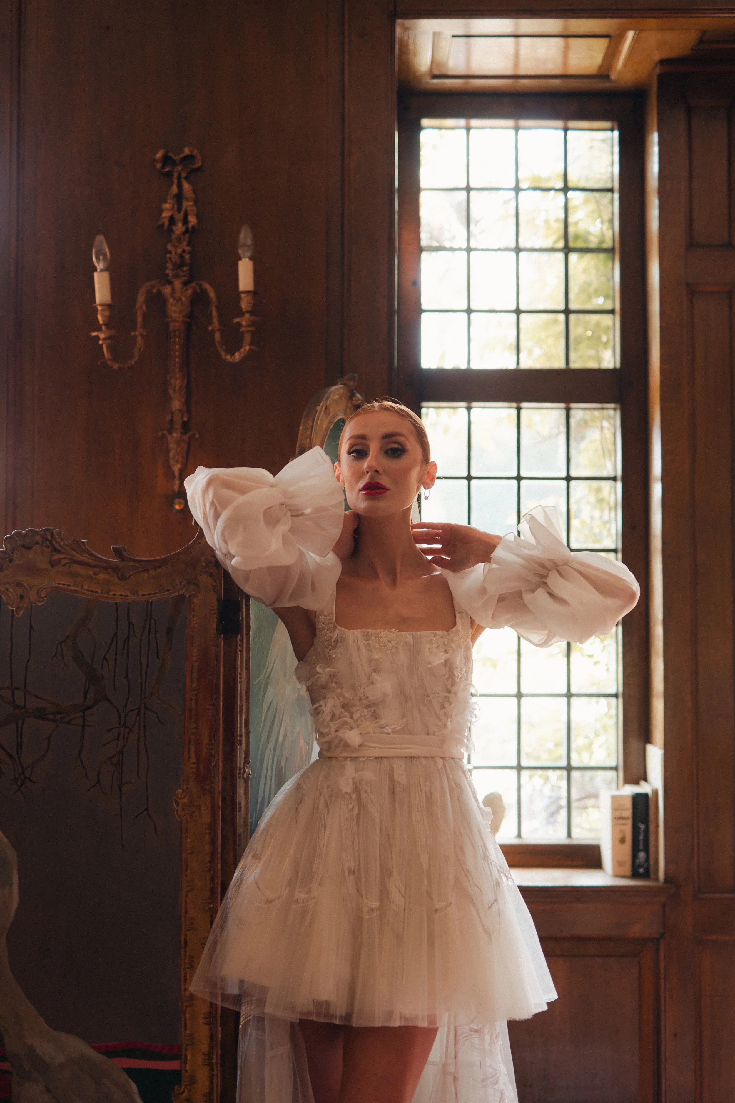 A woman in an elegant white dress with puffed sleeves standing near a mirror and window in a wood-paneled room.