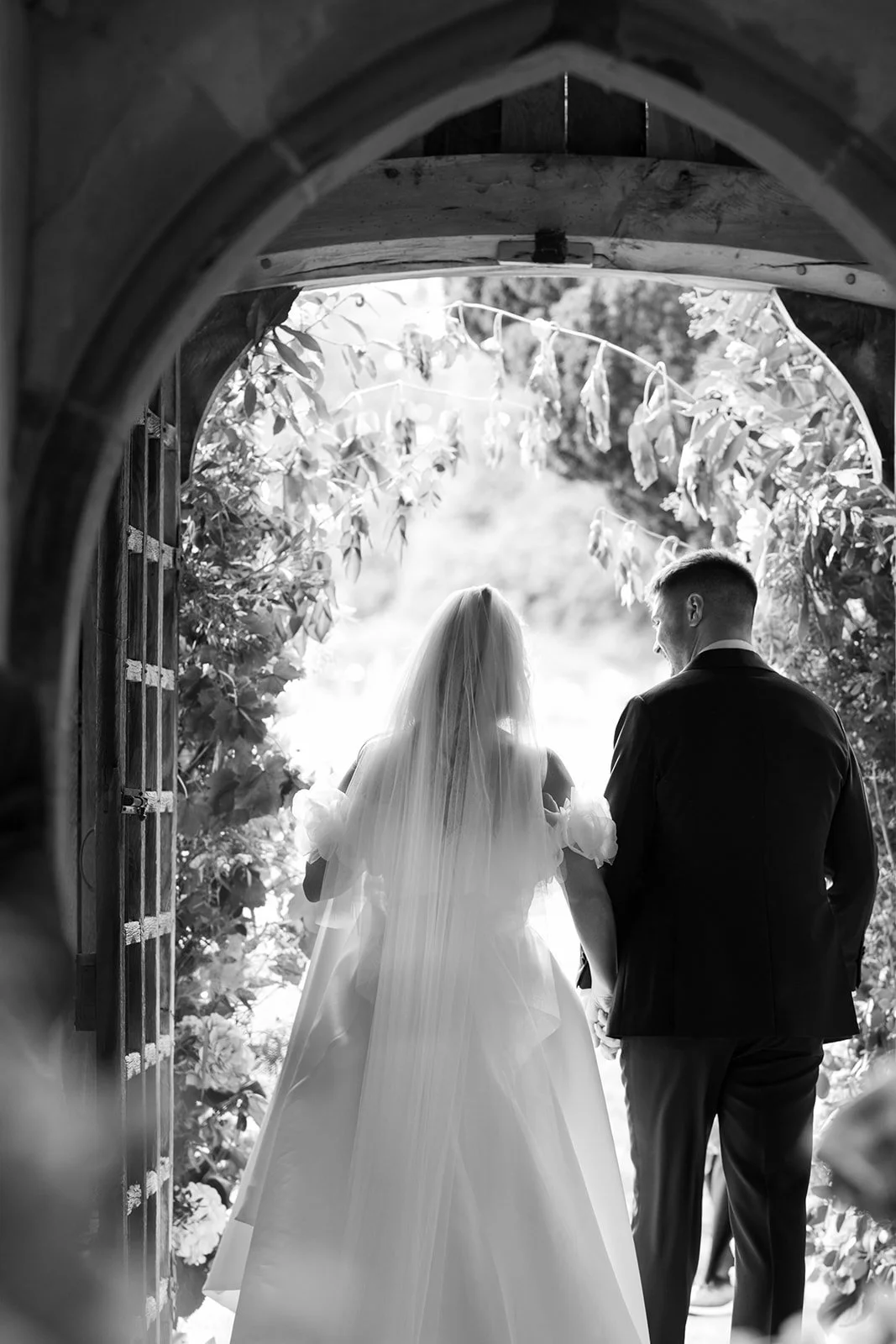 A bride and groom holding hands, walking through an arched garden gate during a wedding, black and white photo.