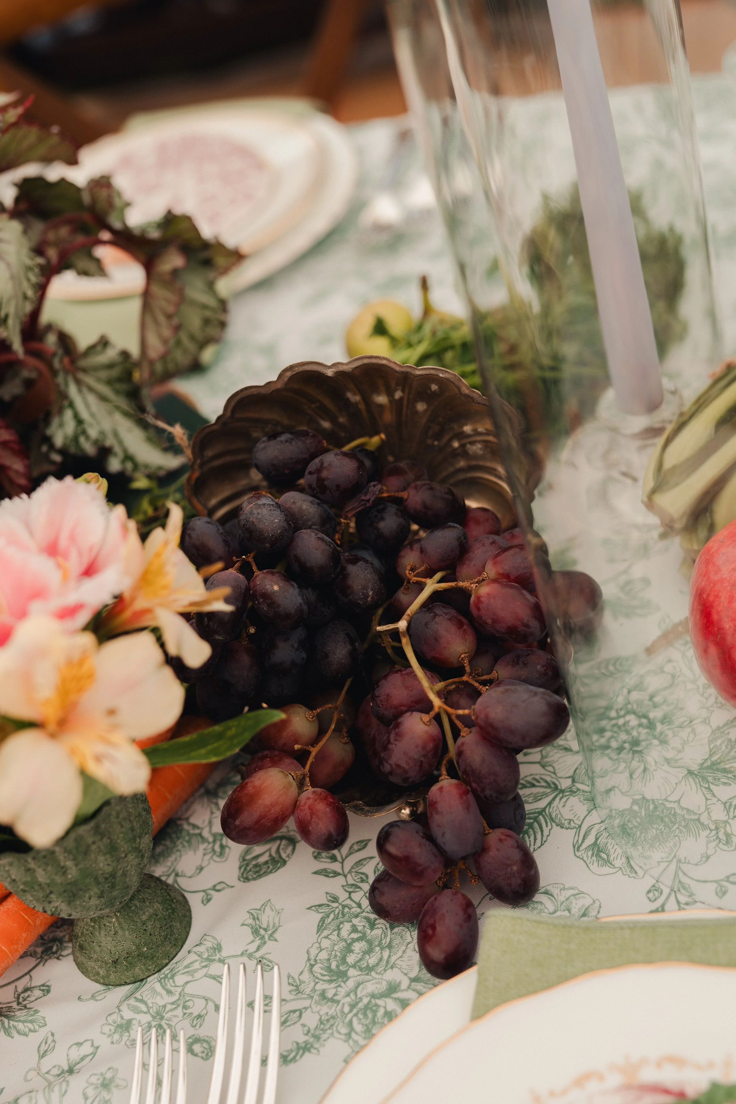 Grapes on a decorative dish on a floral tablecloth with flowers and other tableware nearby.
