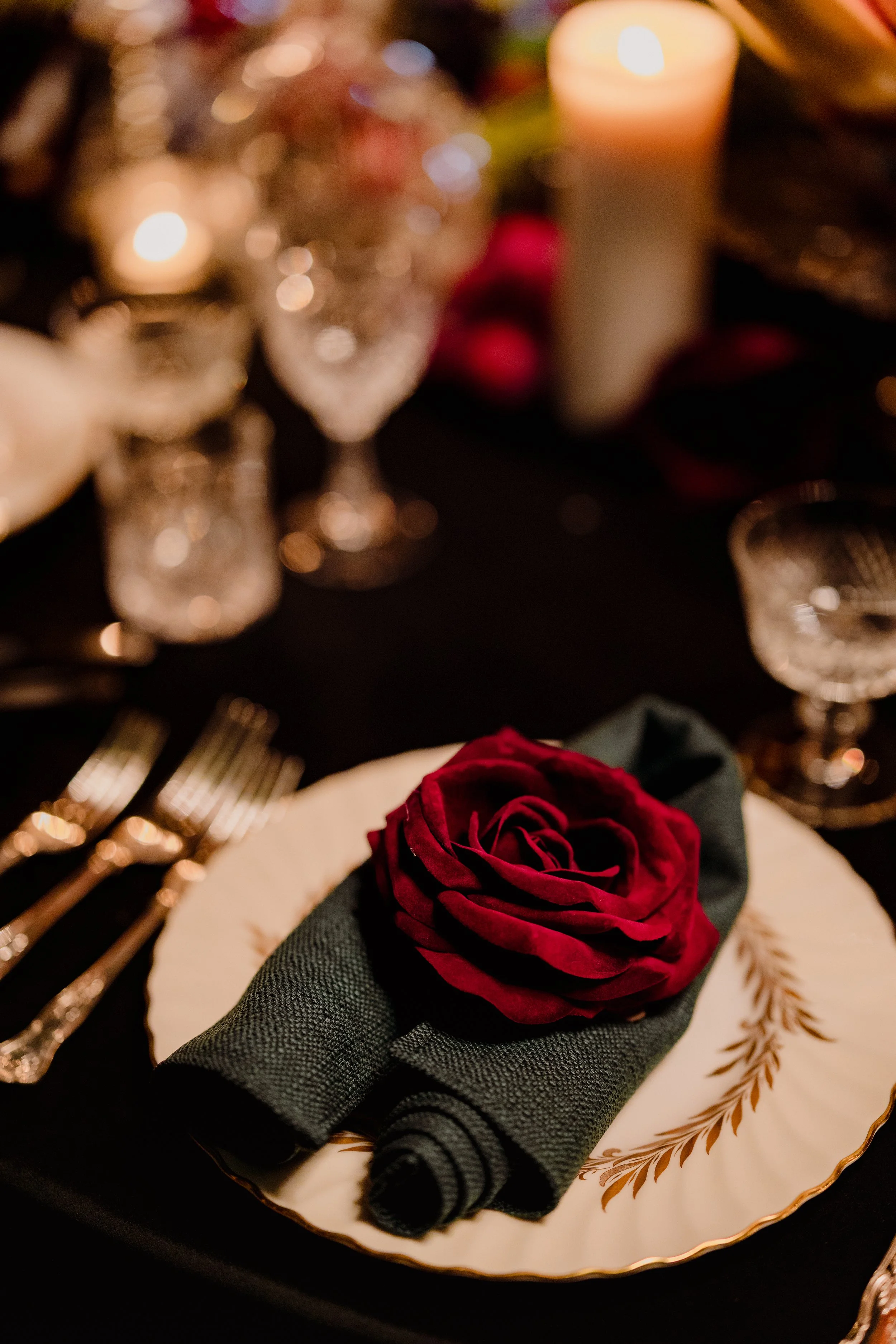 A place setting with a white plate with gold leaf design, black cloth napkin rolled with a dark red rose on top, set on a dark table with silverware and glassware, and blurred candles and decorations in the background.