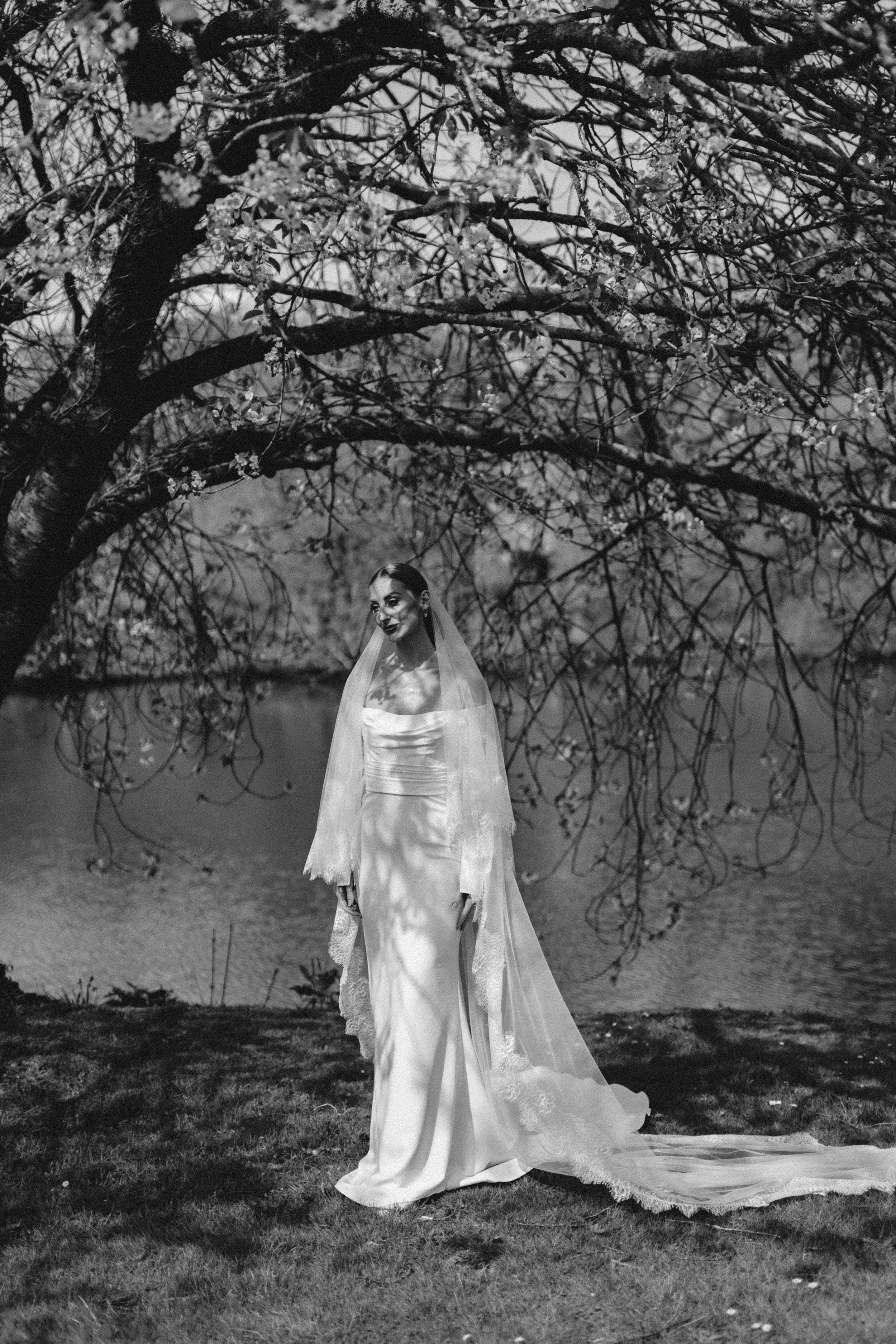 A bride in a white wedding gown stands outdoors beneath a flowering tree, with a lake in the background, captured in black and white.
