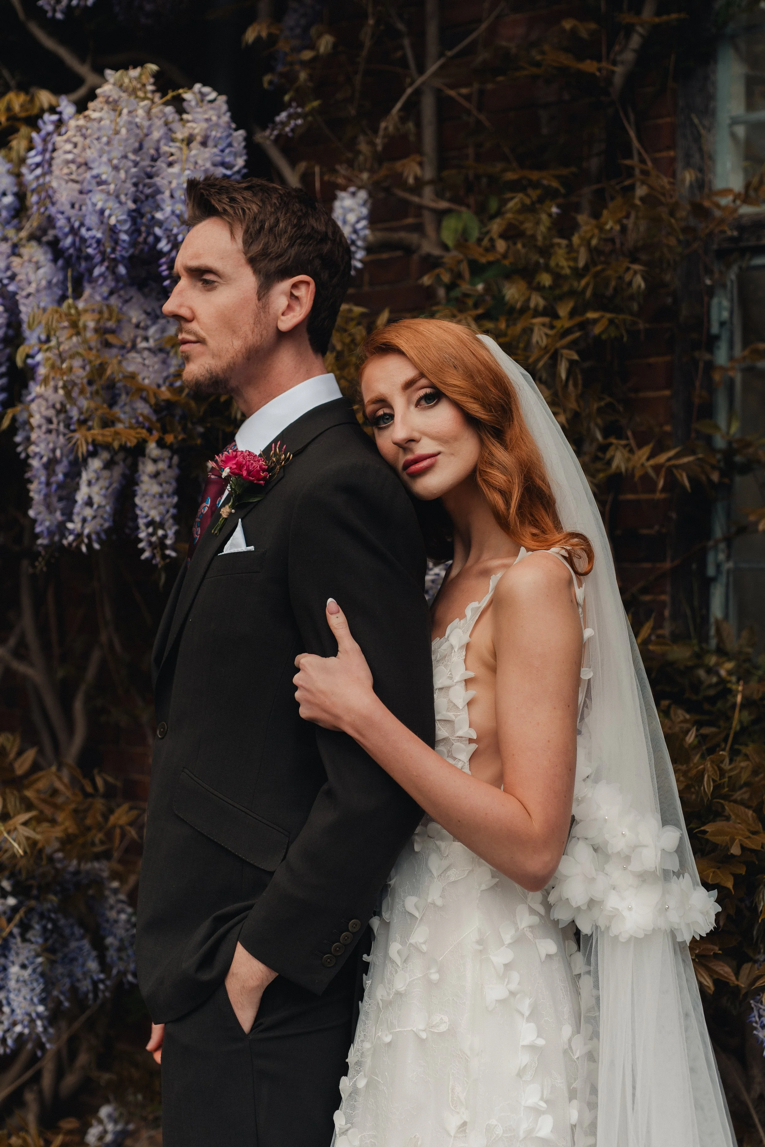A bride with red hair in a white wedding dress holds onto a groom in a black suit and bow tie, standing outdoors with purple flowers and leaves in the background.