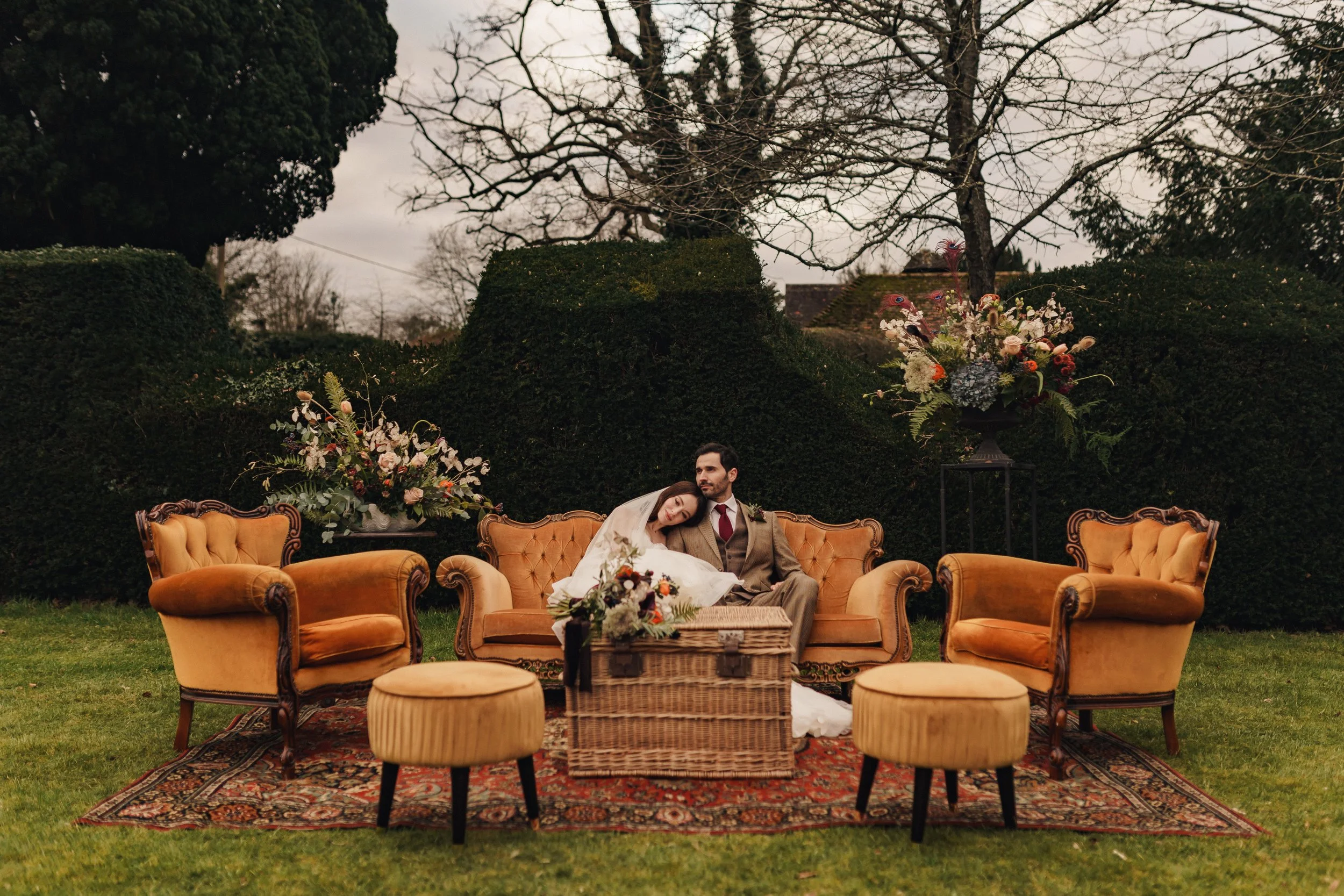 A vintage outdoor wedding scene with a bride and groom sitting on an amber velvet sofa in a lush garden. The bride is resting her head on the groom's shoulder, and they are surrounded by floral arrangements on either side and a woven chest in front of them. Two upholstered stools are positioned in front of the sofa, with a decorative rug underneath on the grass.