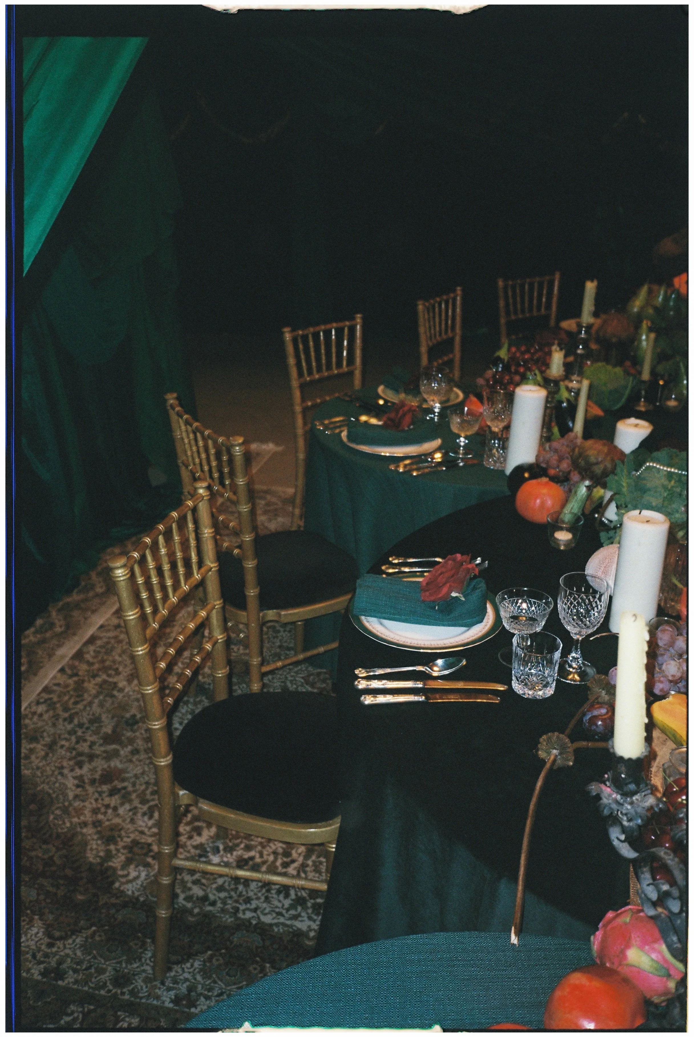 A decorated dining table with gold chairs, set with plates, cutlery, and glasses, surrounded by a dark tablecloth and autumn-themed centerpieces including candles, pumpkins, and grapes.