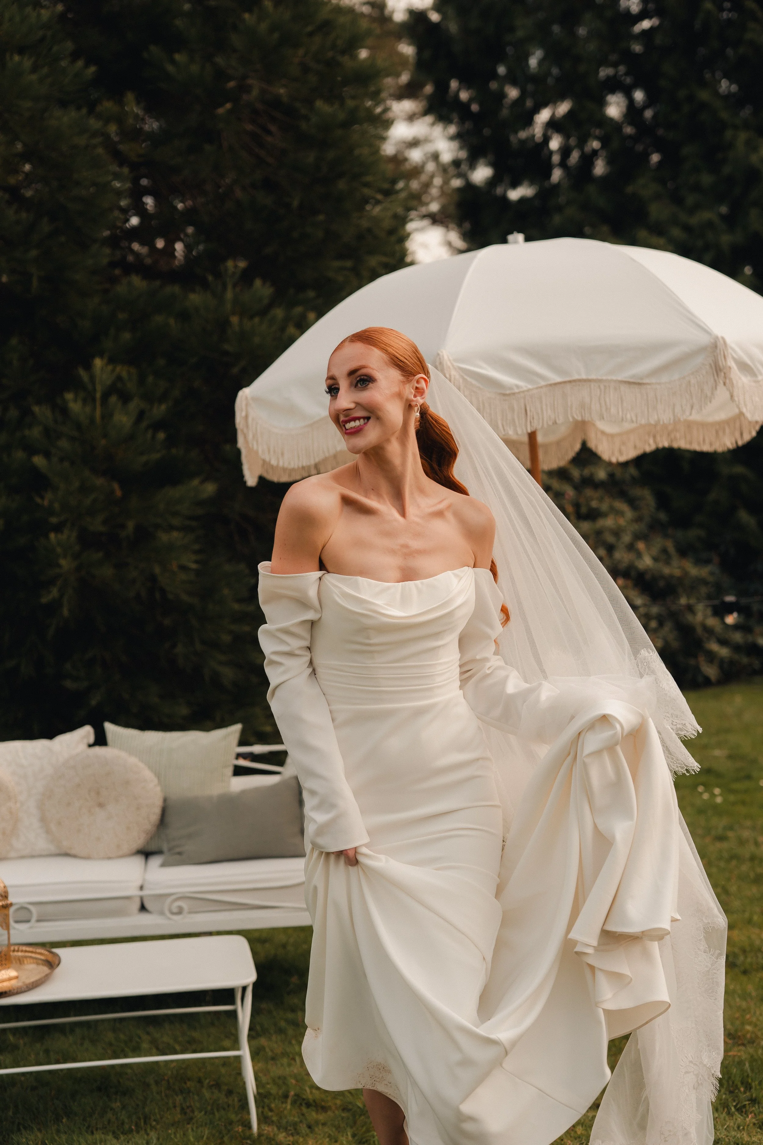 A smiling woman in a white off-shoulder wedding dress holding the skirt, standing outdoors next to a white patio umbrella with fringe, with a grassy area and trees in the background.
