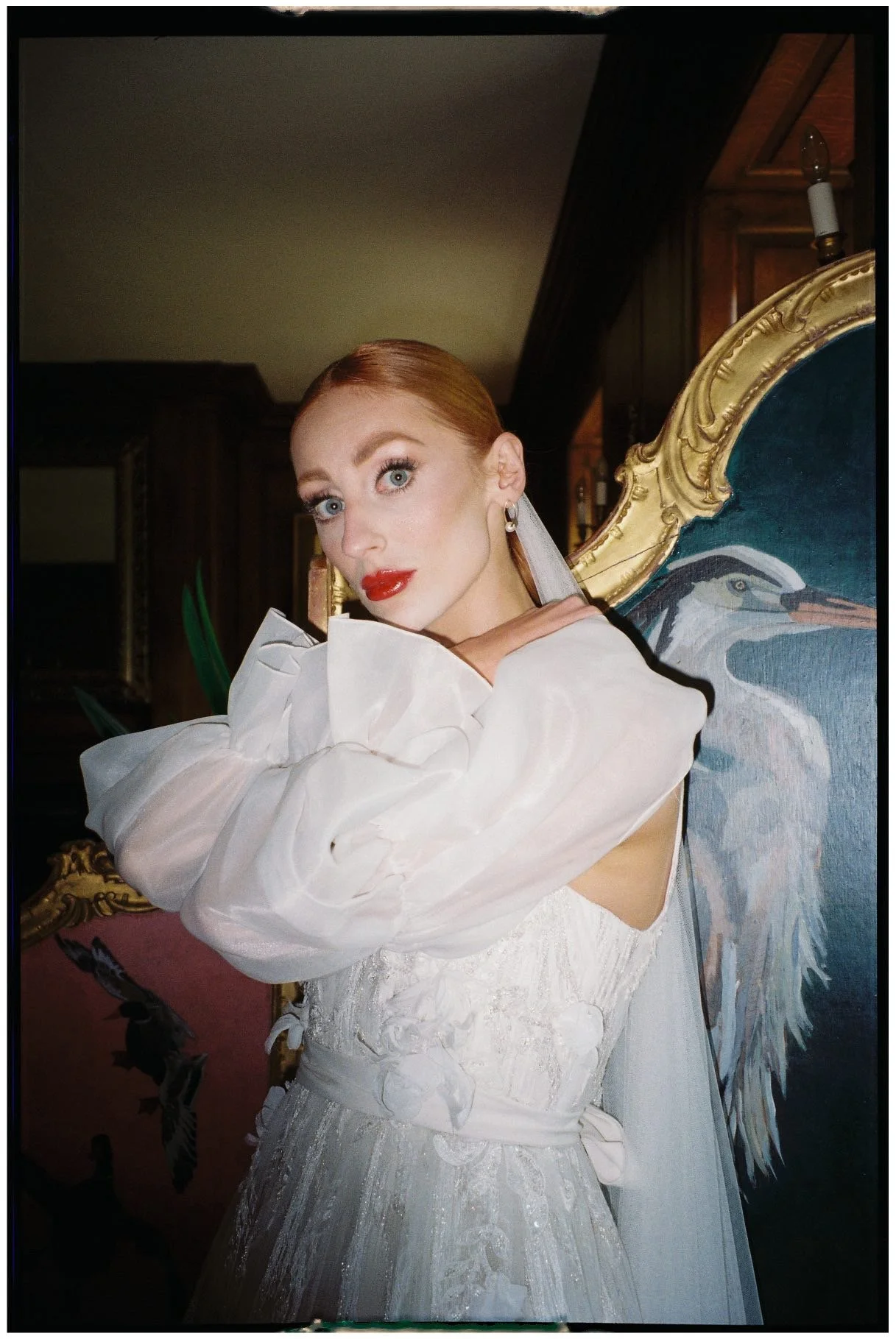 A woman with red hair and blue eyes, wearing a white dress with puffed sleeves, standing in front of a decorative painting of a heron. She has red lipstick, earrings, and a light-colored scarf or veil.