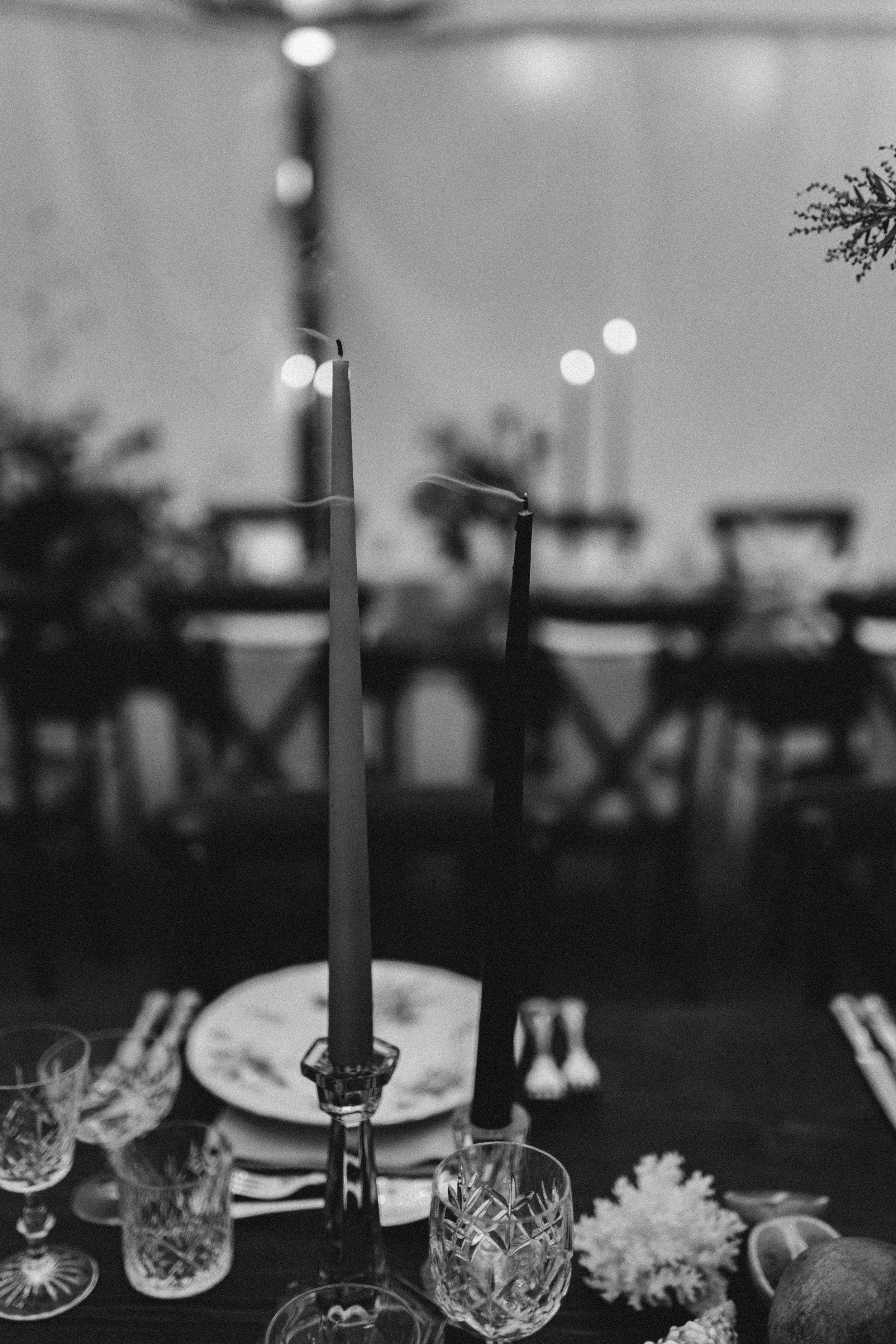 A black and white photo of a dining table with two tall candles, glassware, plates, and a table centerpiece with flowers and a lemon wedge, set for a meal.