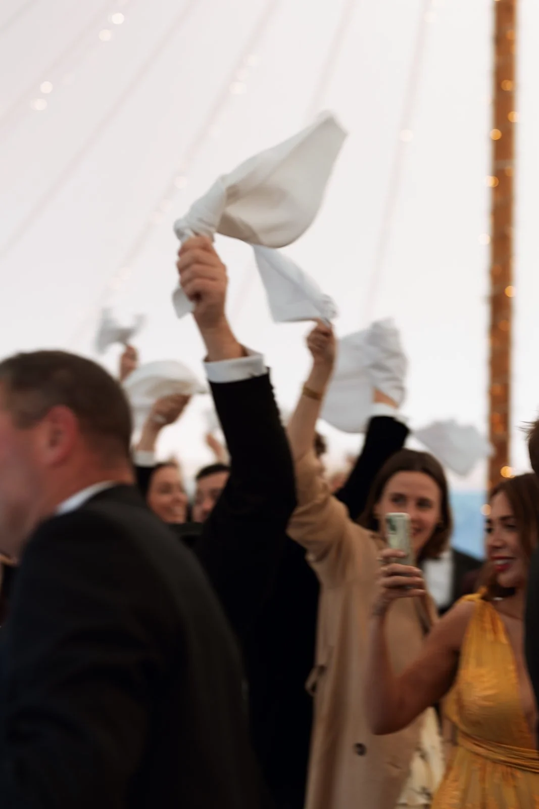 People celebrating at an event, raising white cloth napkins in the air, with some women smiling and using a mobile phone to capture the moment.