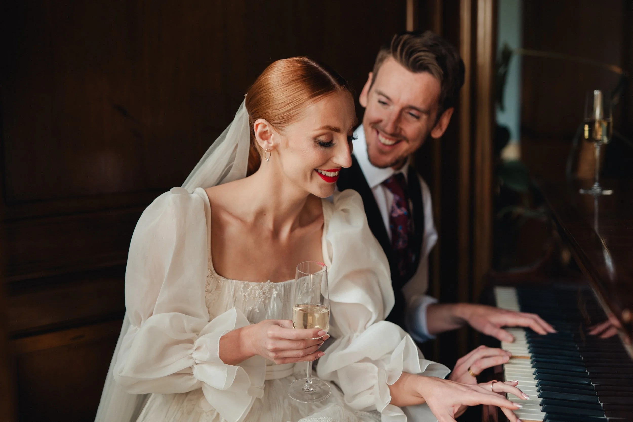 A bride and groom playing piano and smiling, with the bride holding a glass of champagne.