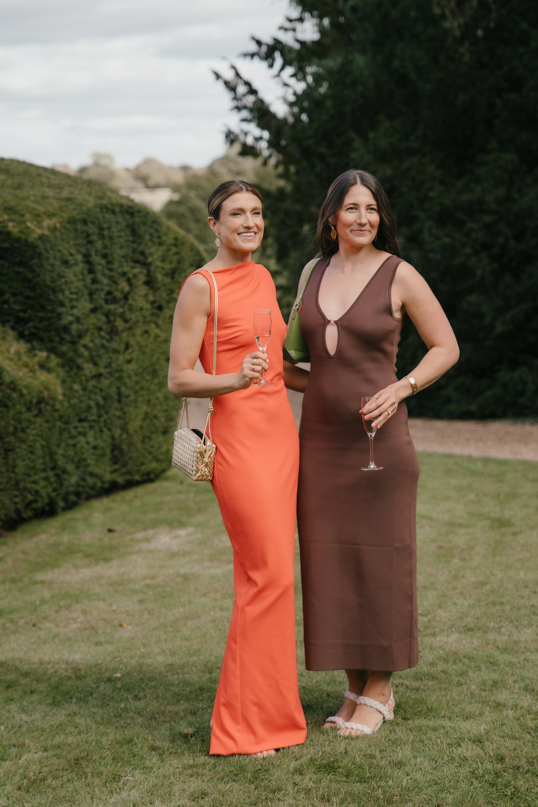 Two women in elegant dresses standing outdoors on grass with bushes and trees in the background, each holding a glass of champagne.
