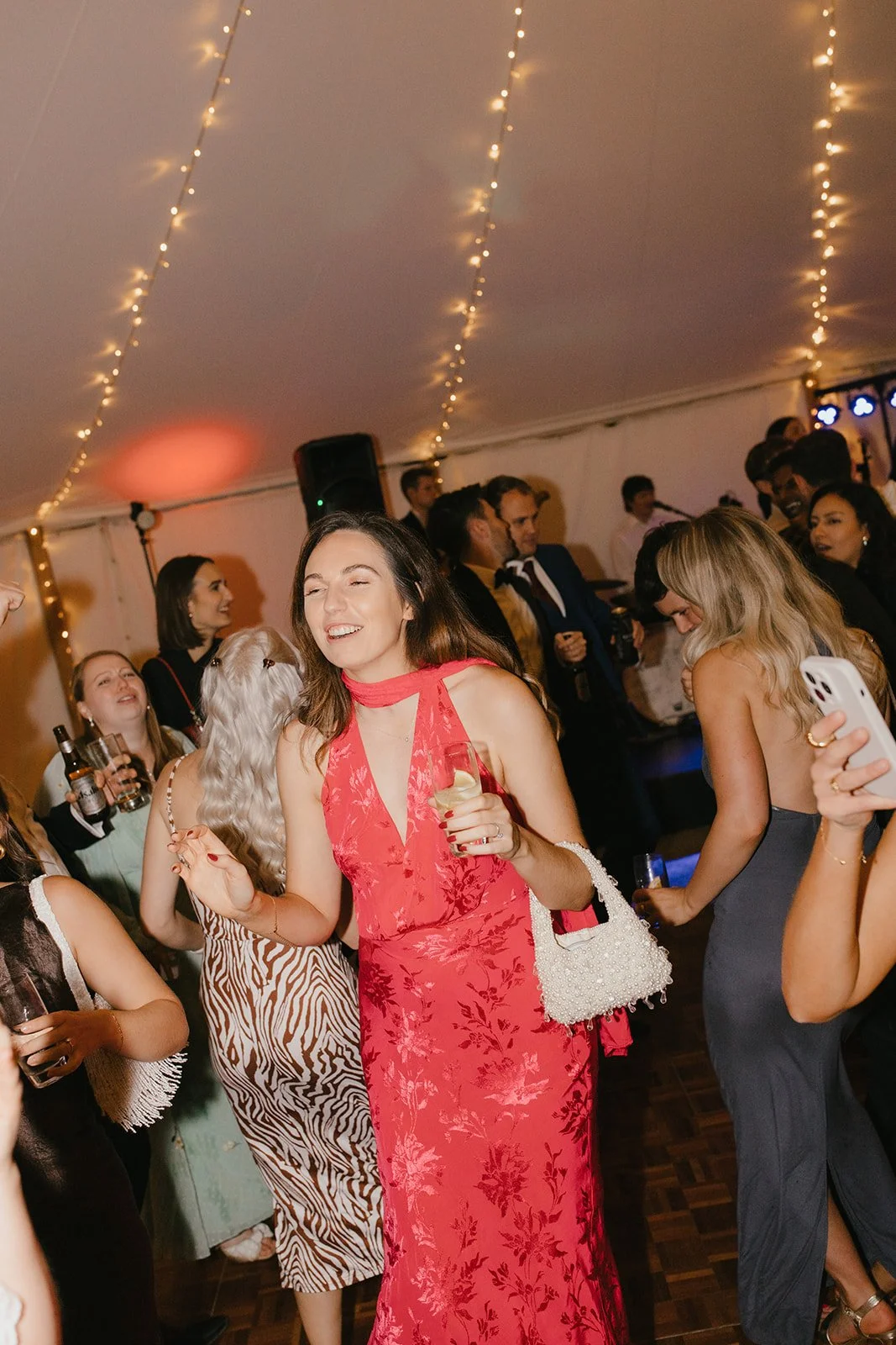 A woman in a red dress dancing and smiling at a party with other guests, some holding drinks, in a decorated tent with string lights.