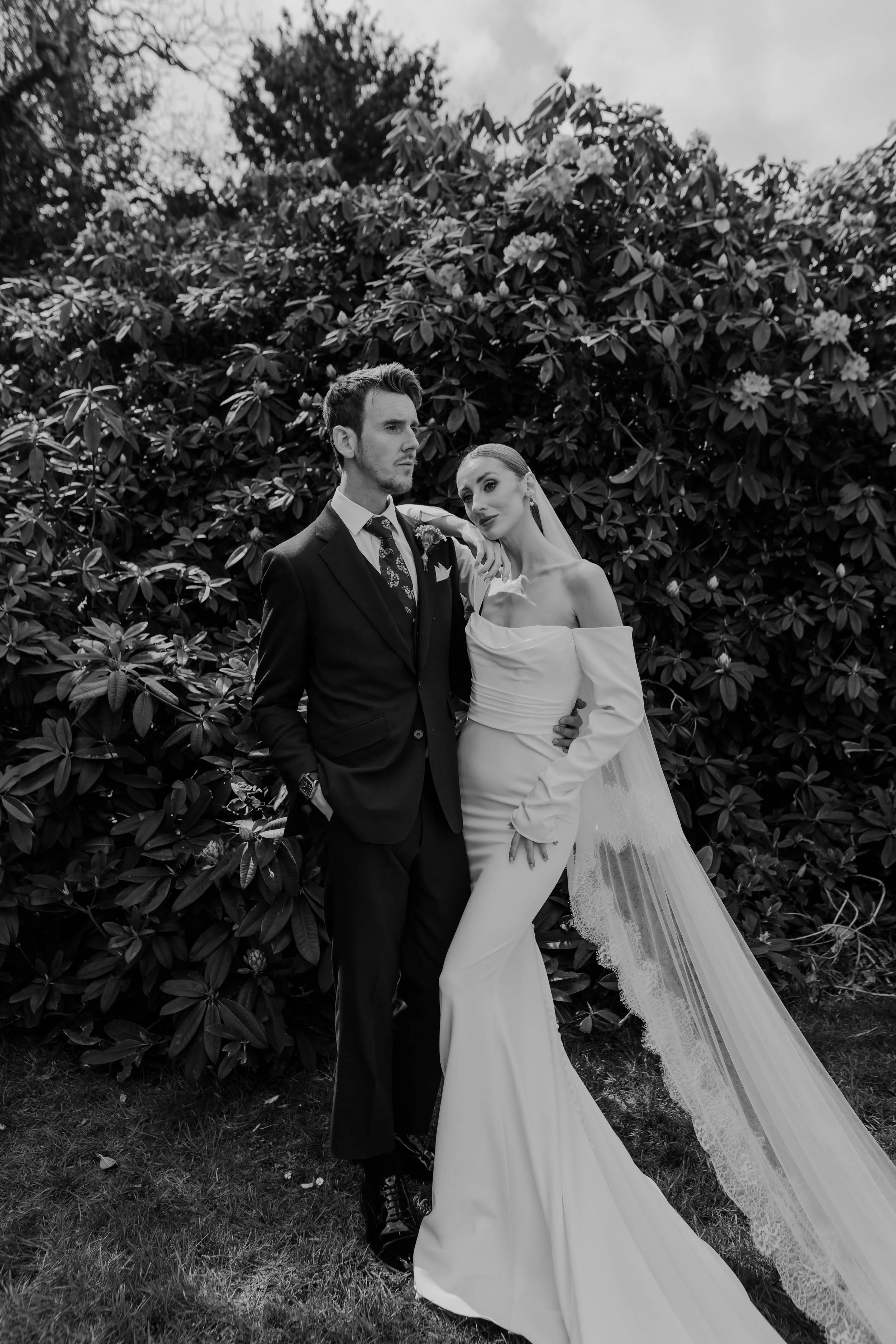 Black and white photo of a bride and groom posing outdoors against a large shrub or bush, with the groom in a suit and the bride in a wedding gown with a veil.