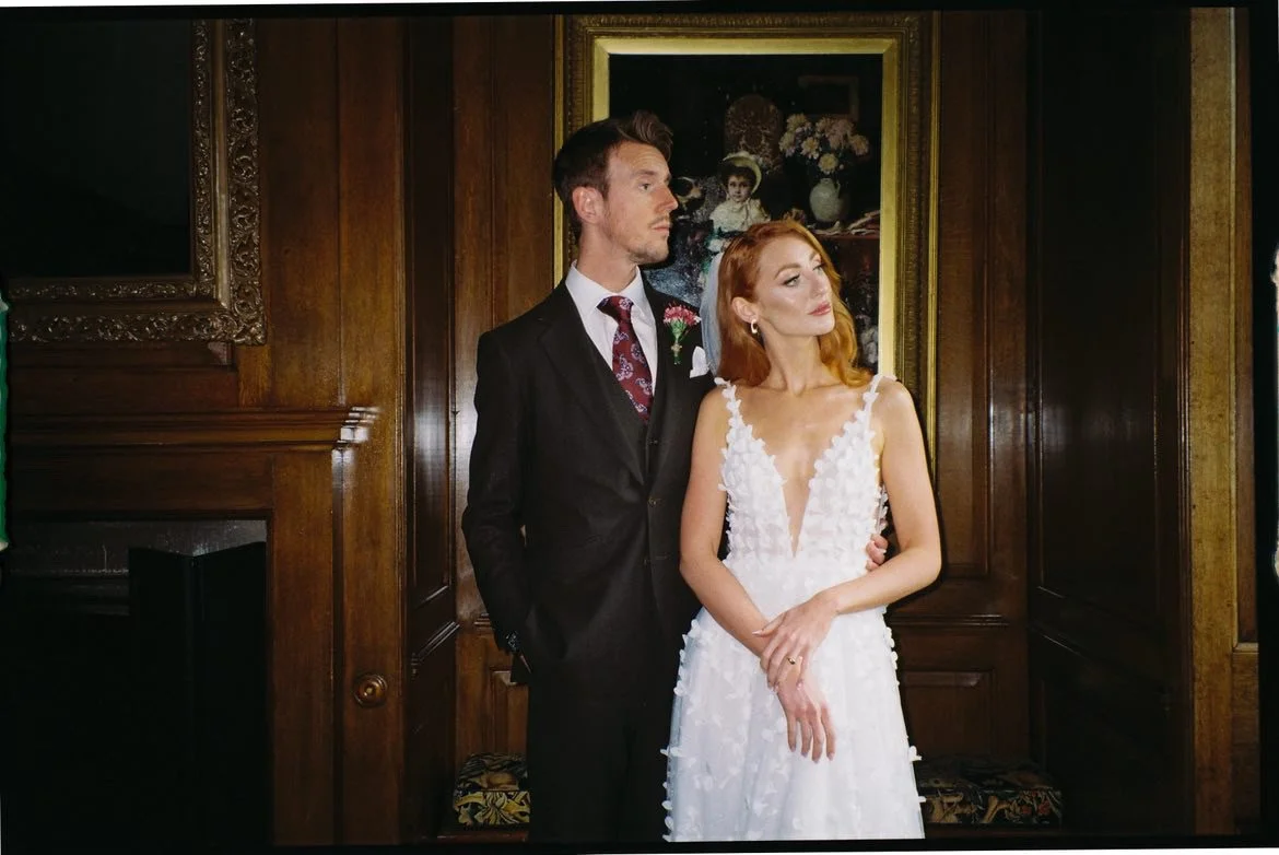 A man in a suit and a woman in a wedding dress standing close together in front of a wooden wall with a framed painting of a girl and flowers.
