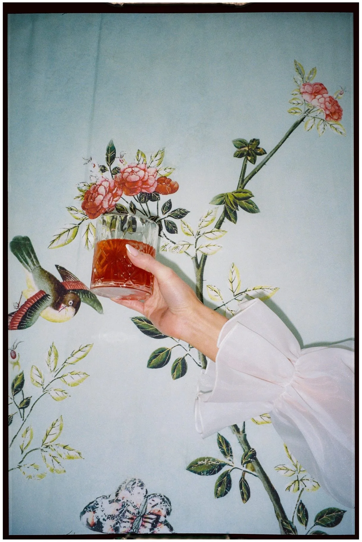 A person's hand holding a glass with a reddish drink in front of a floral wall mural. The person's sleeve is white with a puffy cuff.