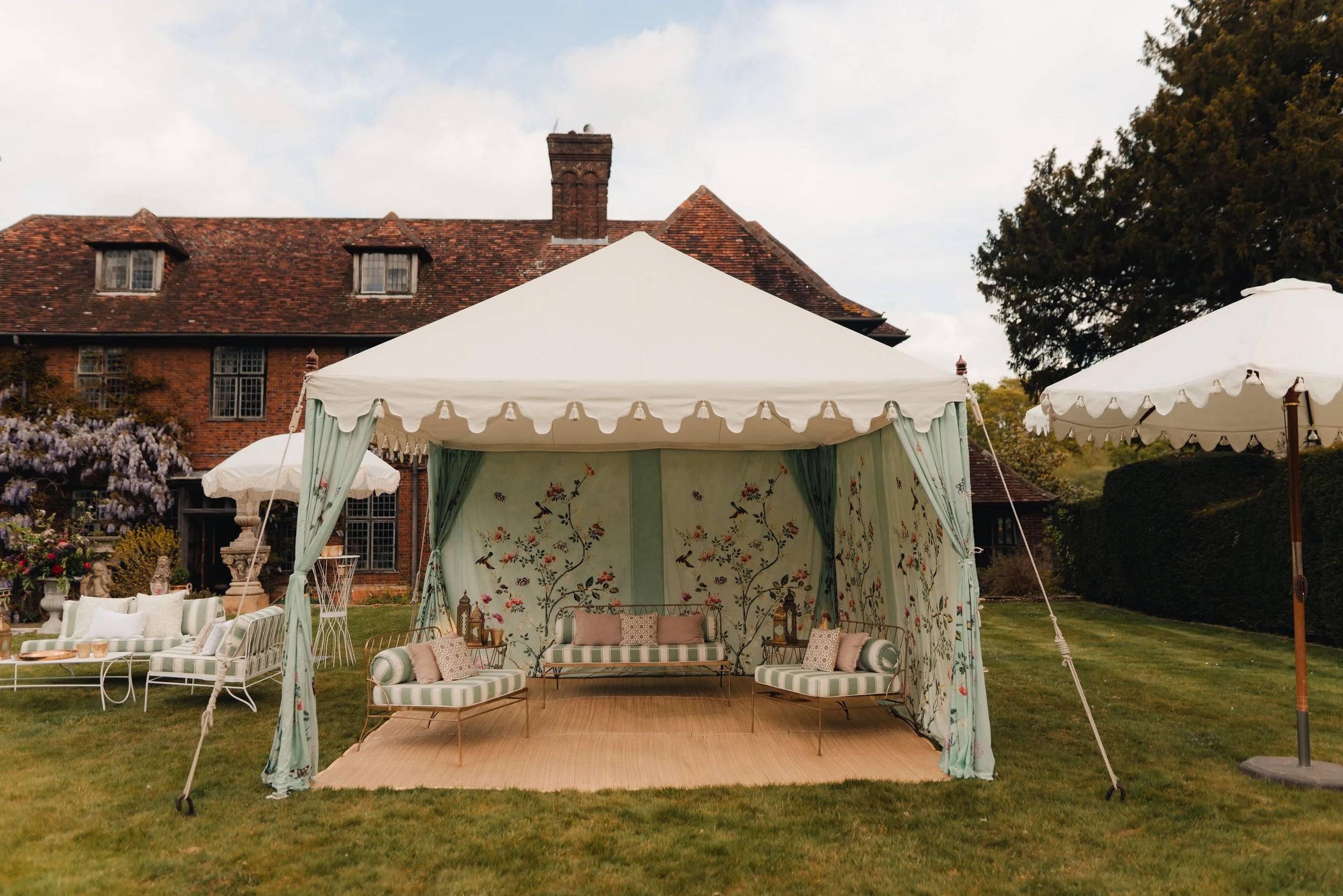 Outdoor seating area under a white canopy tent with floral and bird pattern decor, featuring striped cushions and pillows, set on a grassy lawn in front of a large brick house with trees and umbrellas.