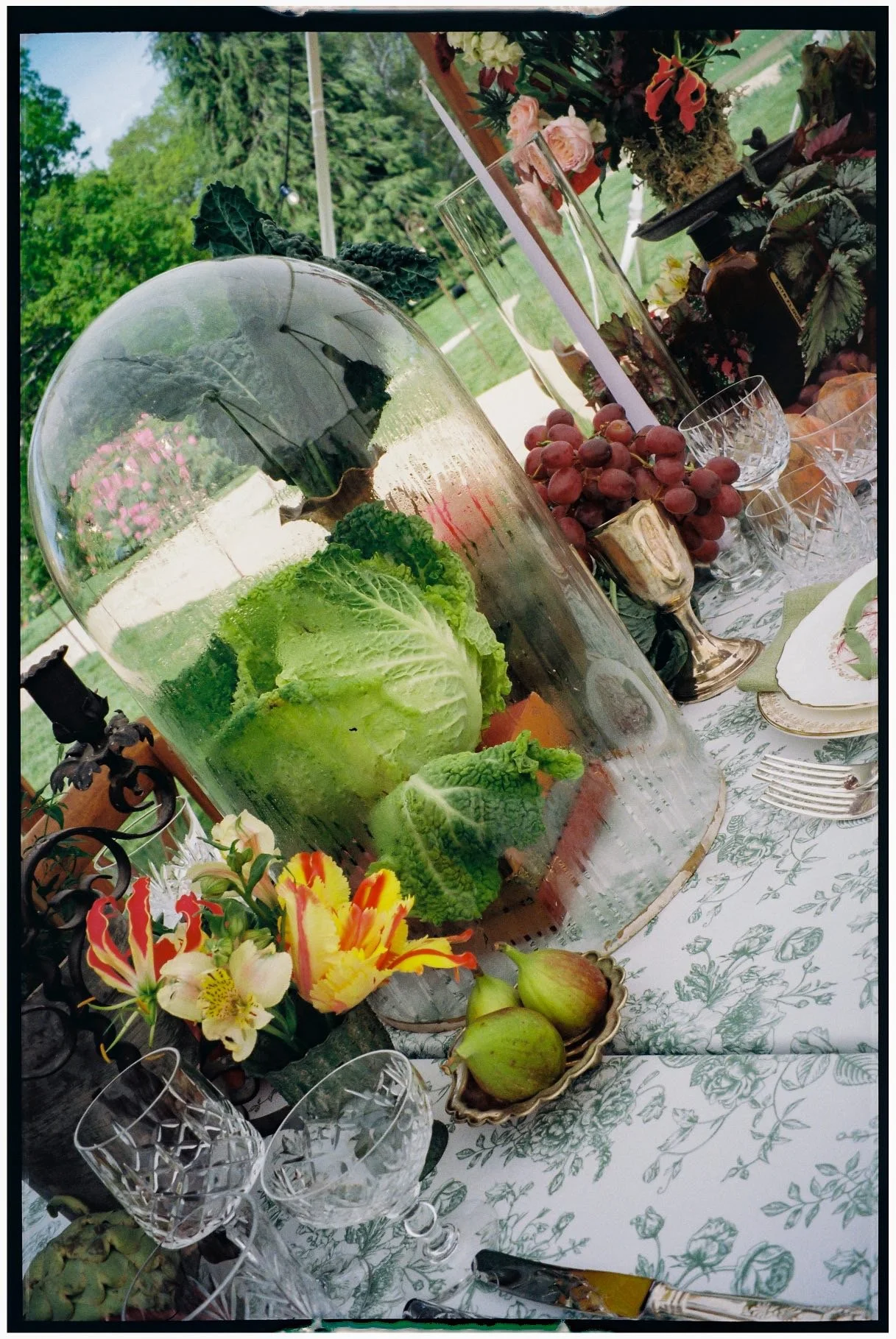 A table with a floral tablecloth, fruit, flowers, and glassware outdoors.
