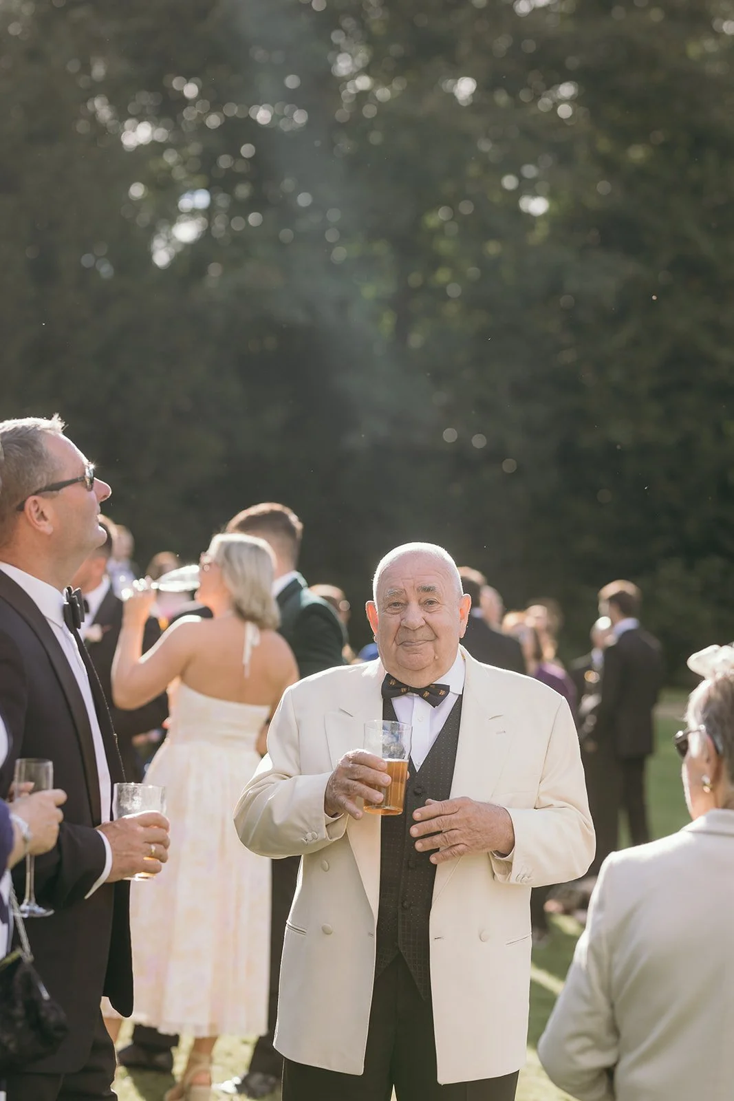 An elderly man in a white tuxedo with a black bow tie holding a glass of beer at an outdoor wedding reception.