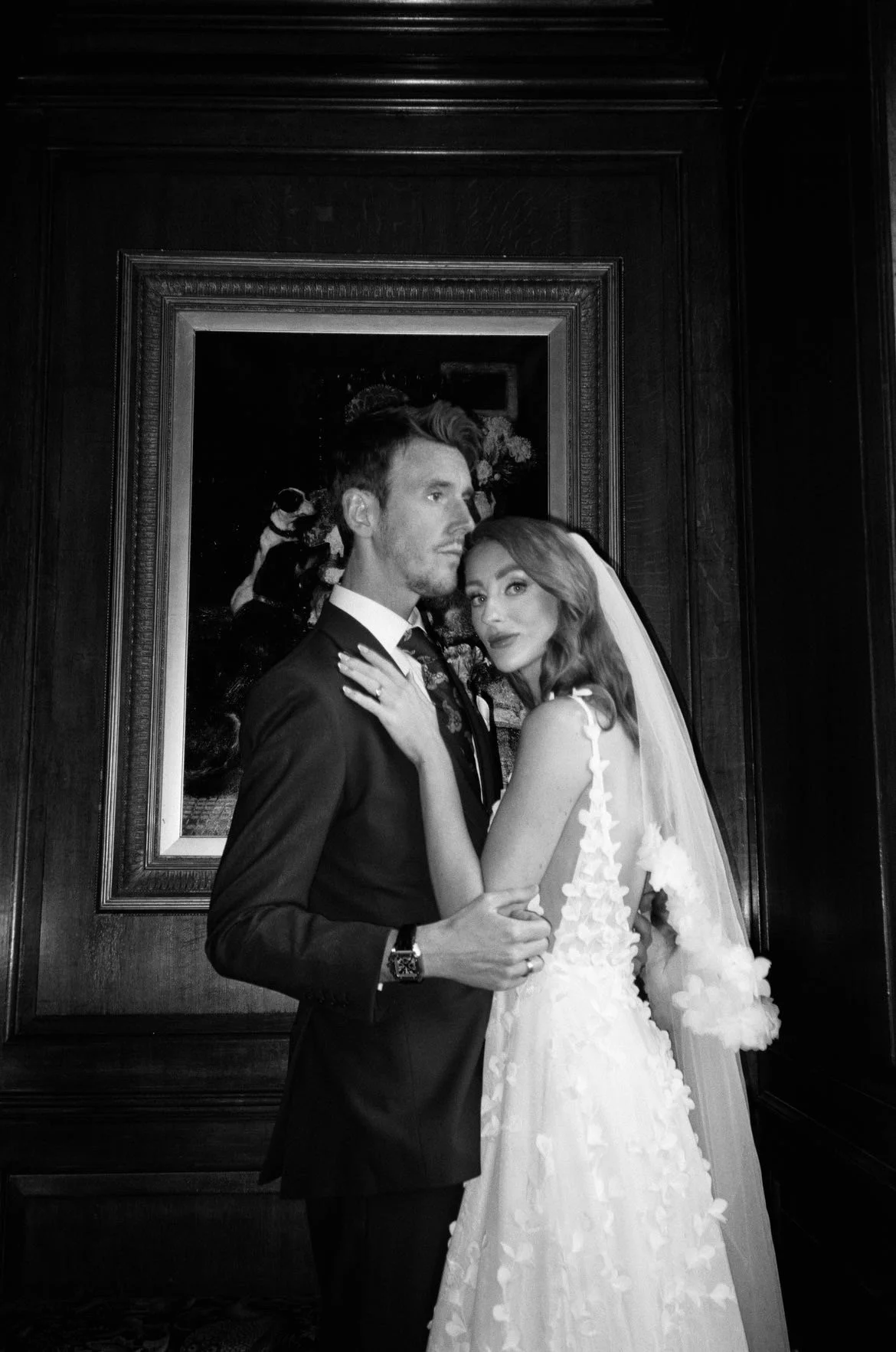 Black and white photo of a bride and groom posing in front of a painting. The bride has long hair, a veil, and a gown with floral details. The groom has short hair, a beard, and is wearing a suit. They are embracing each other indoors.