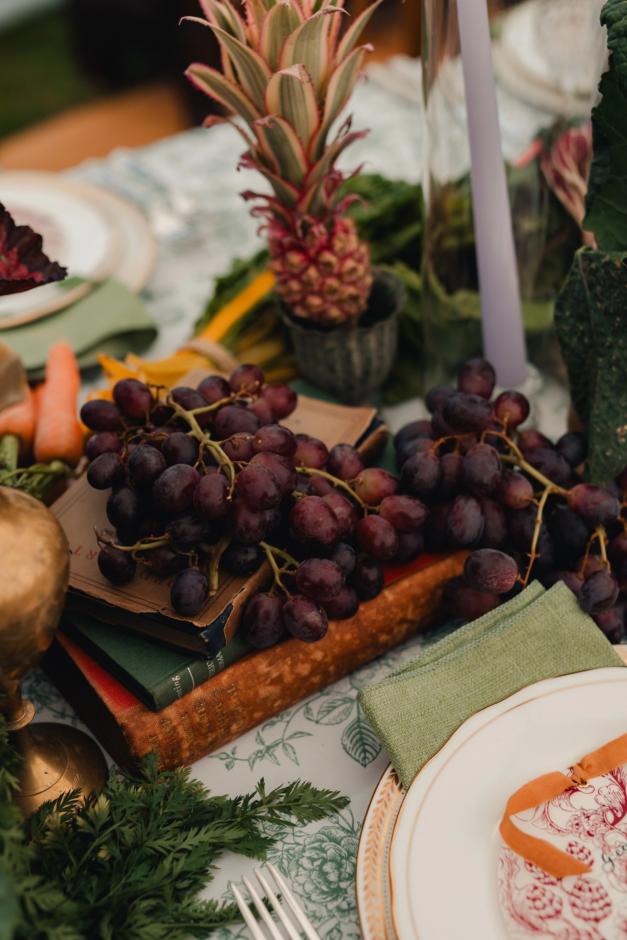 A table setting with a bunch of red grapes on top of magazines, surrounded by pineapple, carrots, greenery, and a decorative plate with an orange napkin.