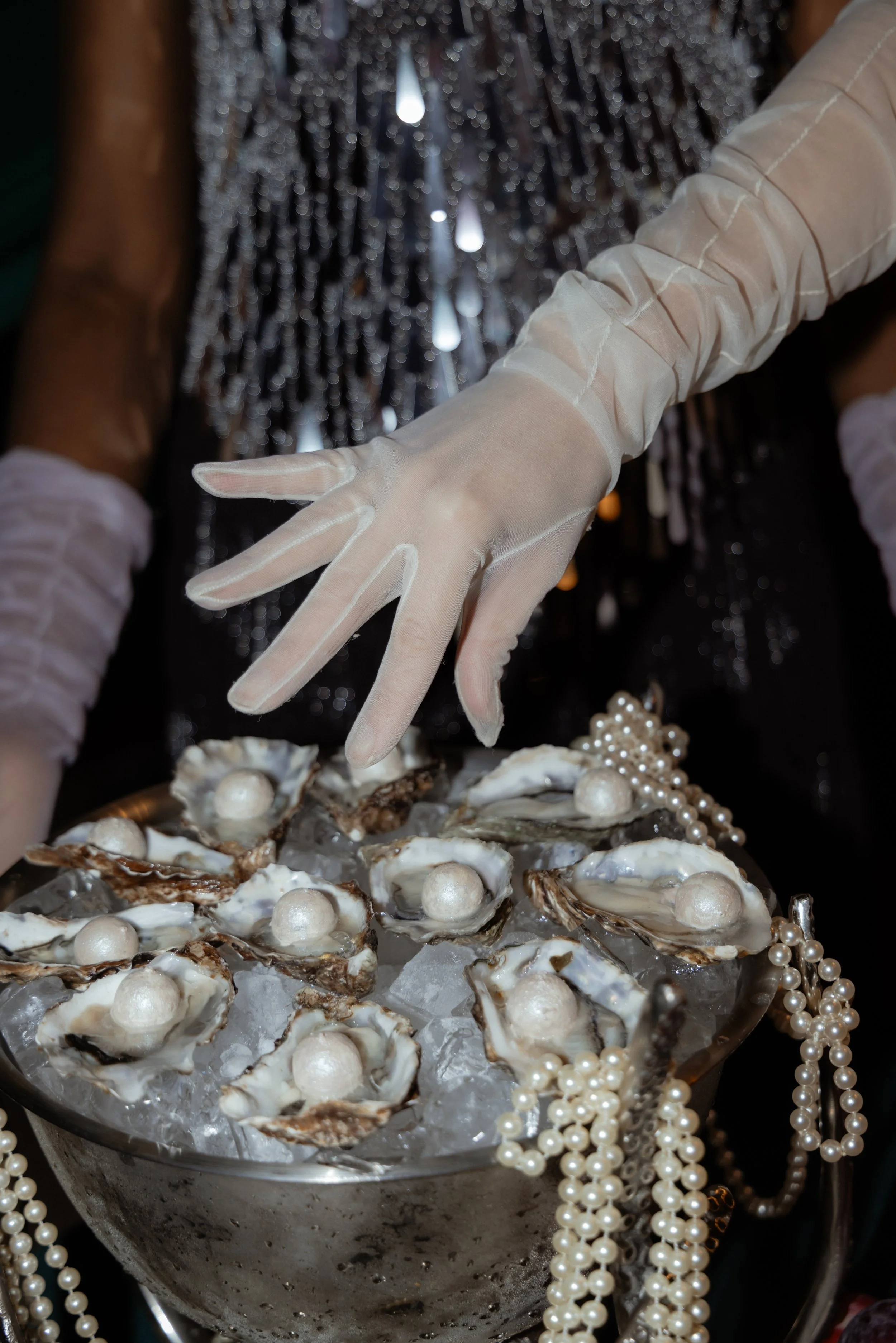Close-up of a person's stacked hands wearing long white gloves, over a tray of open oysters on ice, decorated with pearls and a pearl necklace, with a black sequined background.