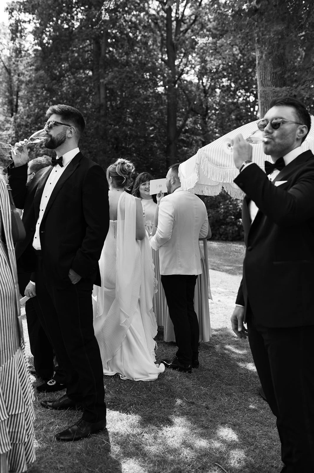 Black and white photo of a wedding celebration outdoors with guests in formal attire, including tuxedos and a bridal gown, mingling and drinking wine under trees.