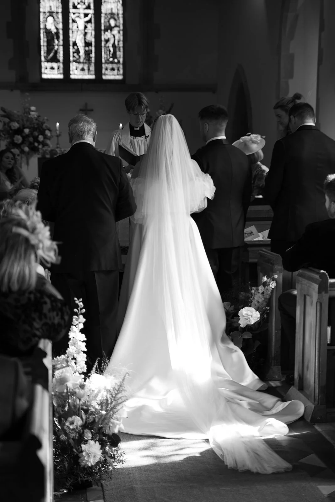 Wedding ceremony inside a church with bride in a long white gown with veil and four men in suits, officiant, stained glass windows, floral arrangements, and guests seated.