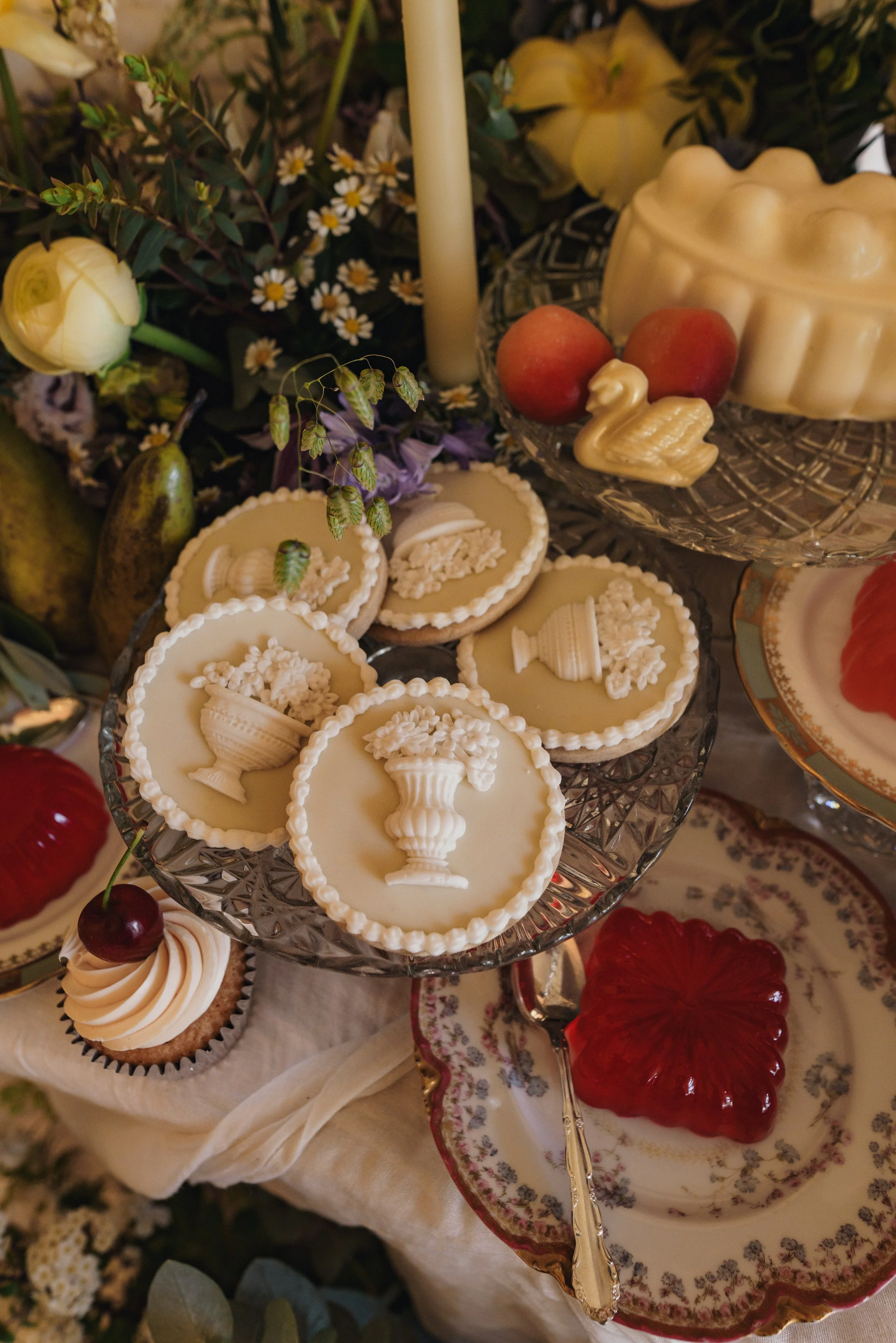 A dessert table featuring white chocolate medallions with urn and floral motifs, a cupcake with a cherry on top, red jelly dessert, and various fruits, surrounded by flowers and a candle.
