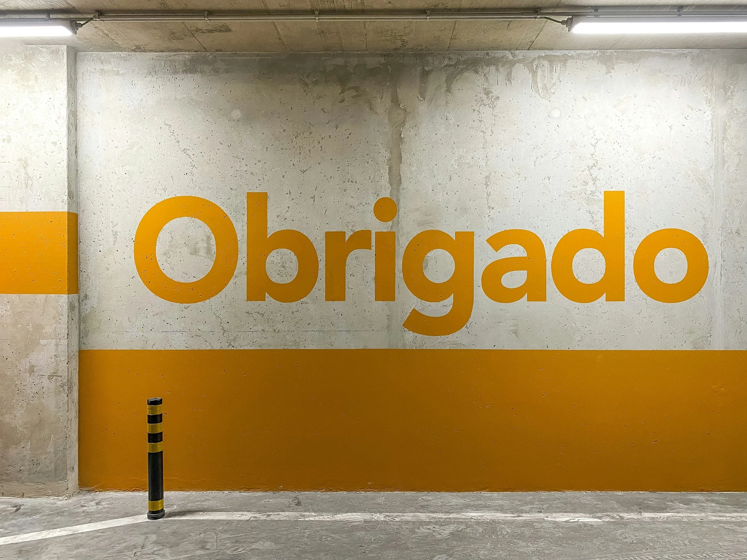 A concrete wall in a carpark with the word 'Obrigado', thankyou in portuguese, painted in large orange letters, with orange bands at the top and bottom of the wall. There is a black and yellow striped pole at the bottom.