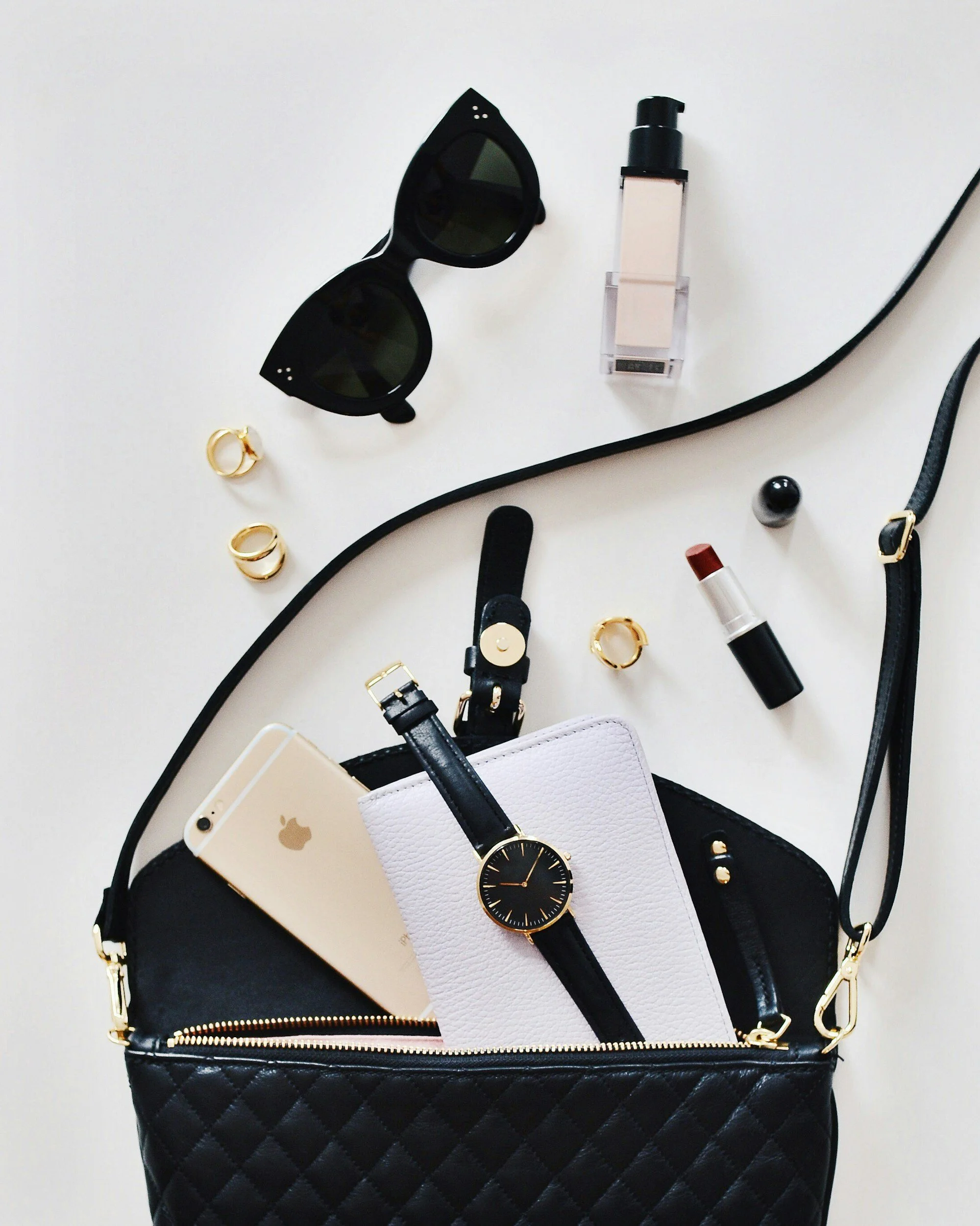 Black quilted handbag with gold zipper, containing a white tablet, a black wristwatch, and a white phone. Items scattered around include black sunglasses, several gold rings, a bottle of foundation, lipstick, and small black and silver accessories.
