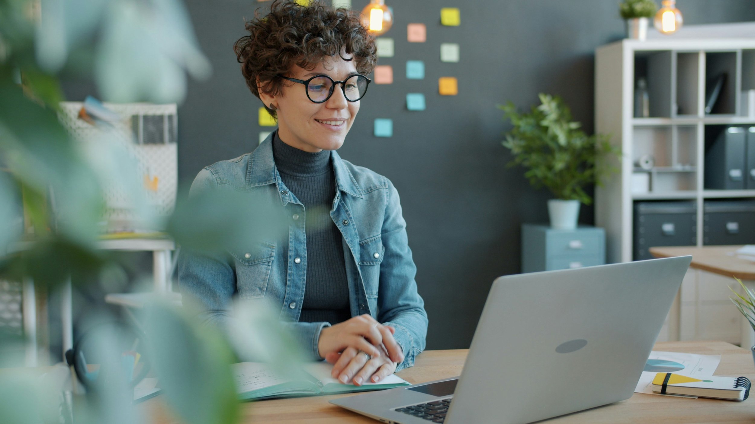 Woman with curly hair and glasses working at a desk with a laptop, notebooks, and papers, preparing for a coaching session  with Your Coach Terri from UnSplash.