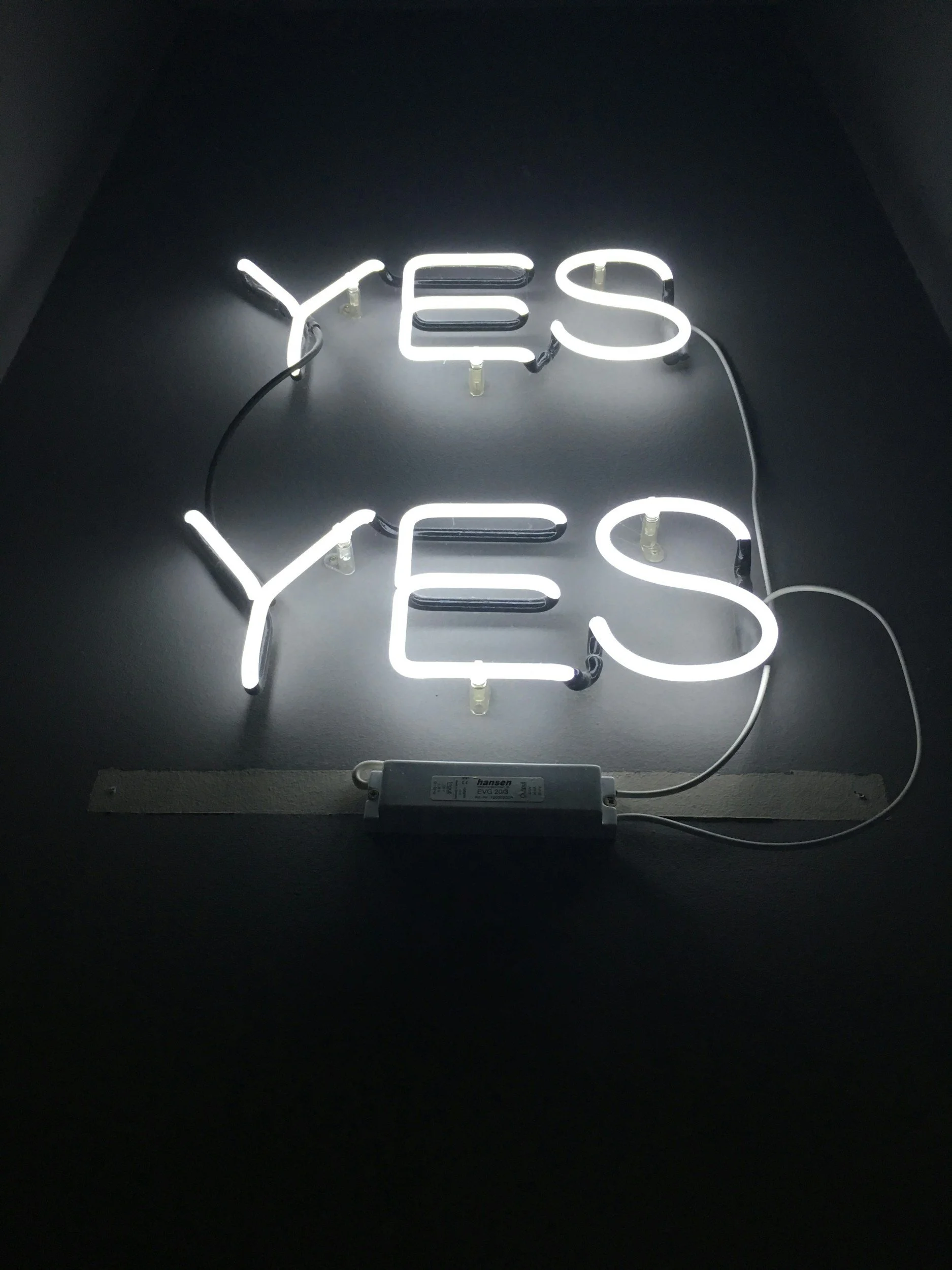 A neon sign displaying the words 'YES' and 'YEs' in white light, mounted on a black wall with visible electrical wires.