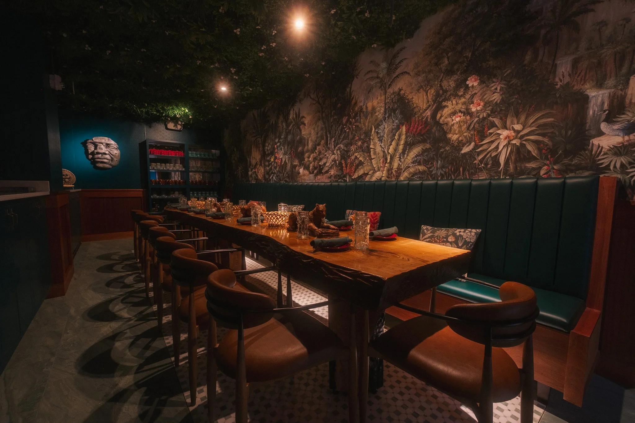 A dimly lit restaurant or dining room with a long wooden table, navy blue upholstered bench, and brown chairs. The wall features a large tropical jungle mural with plants and flowers, and a mask or sculpture of a face is mounted on a dark green wall.