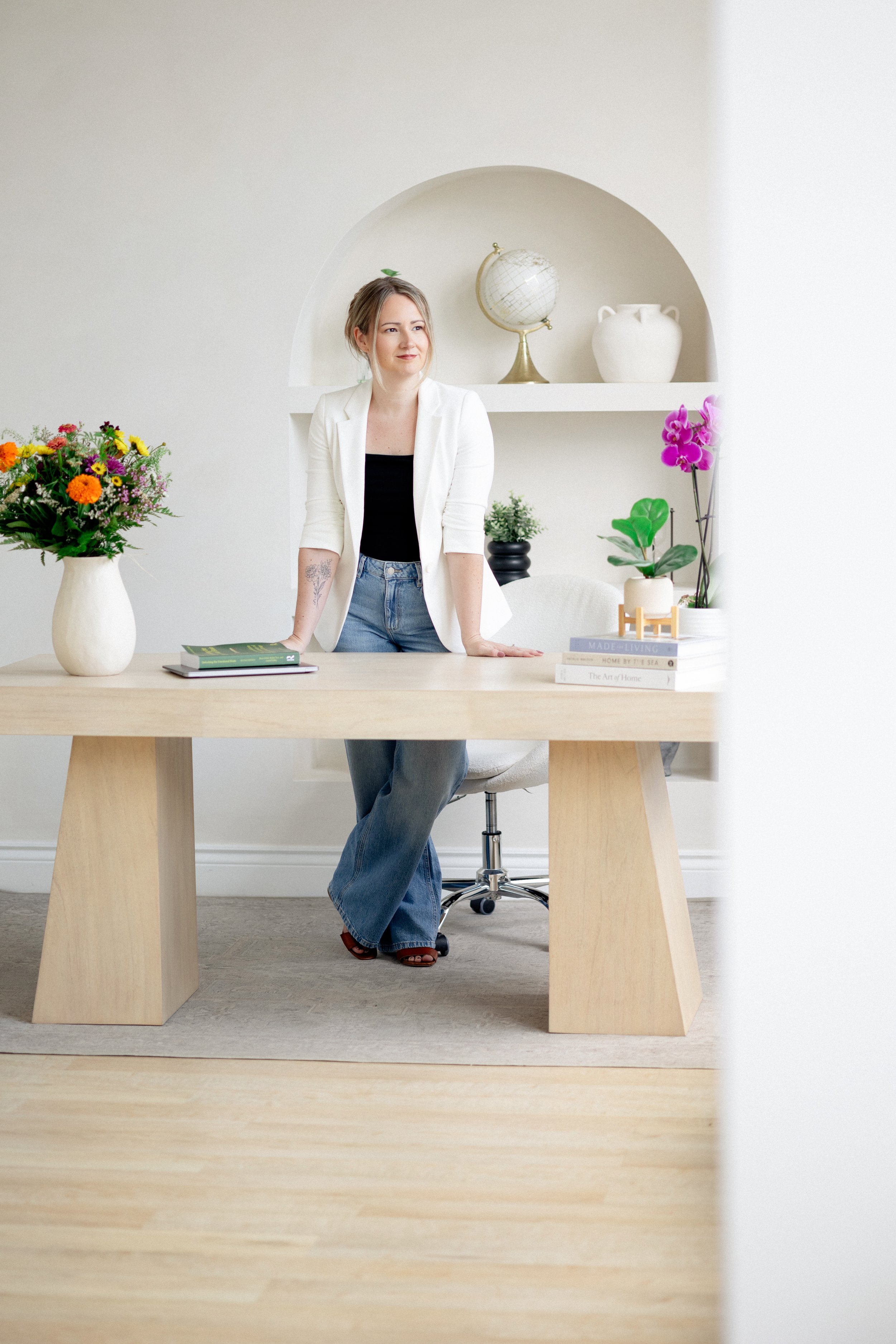 A woman standing behind a wooden desk in a bright room with minimal decor, flower arrangements, and books.