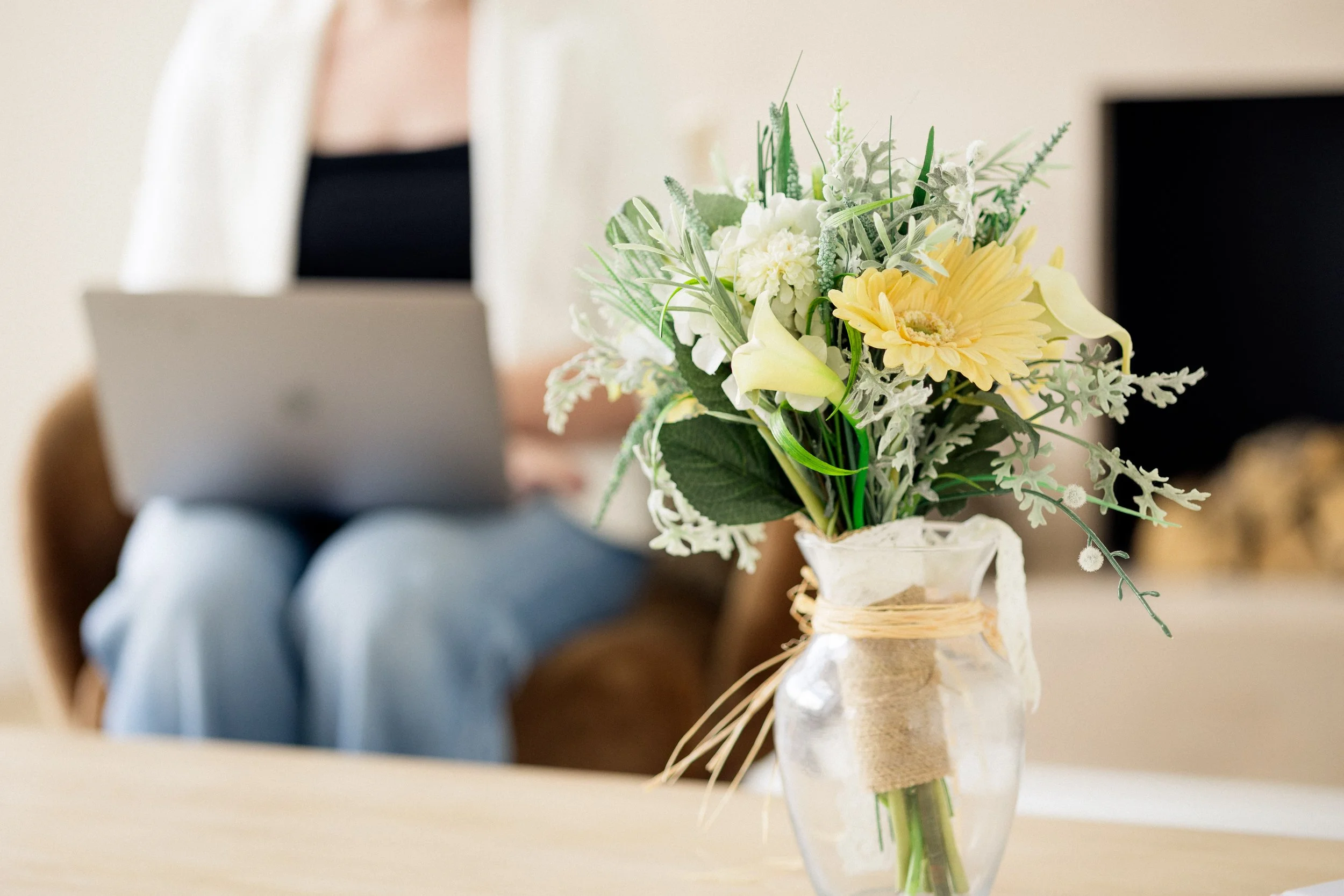 Close-up of a glass vase with a bouquet of white and yellow flowers on a wooden table, with a person sitting on a sofa using a laptop in the background.