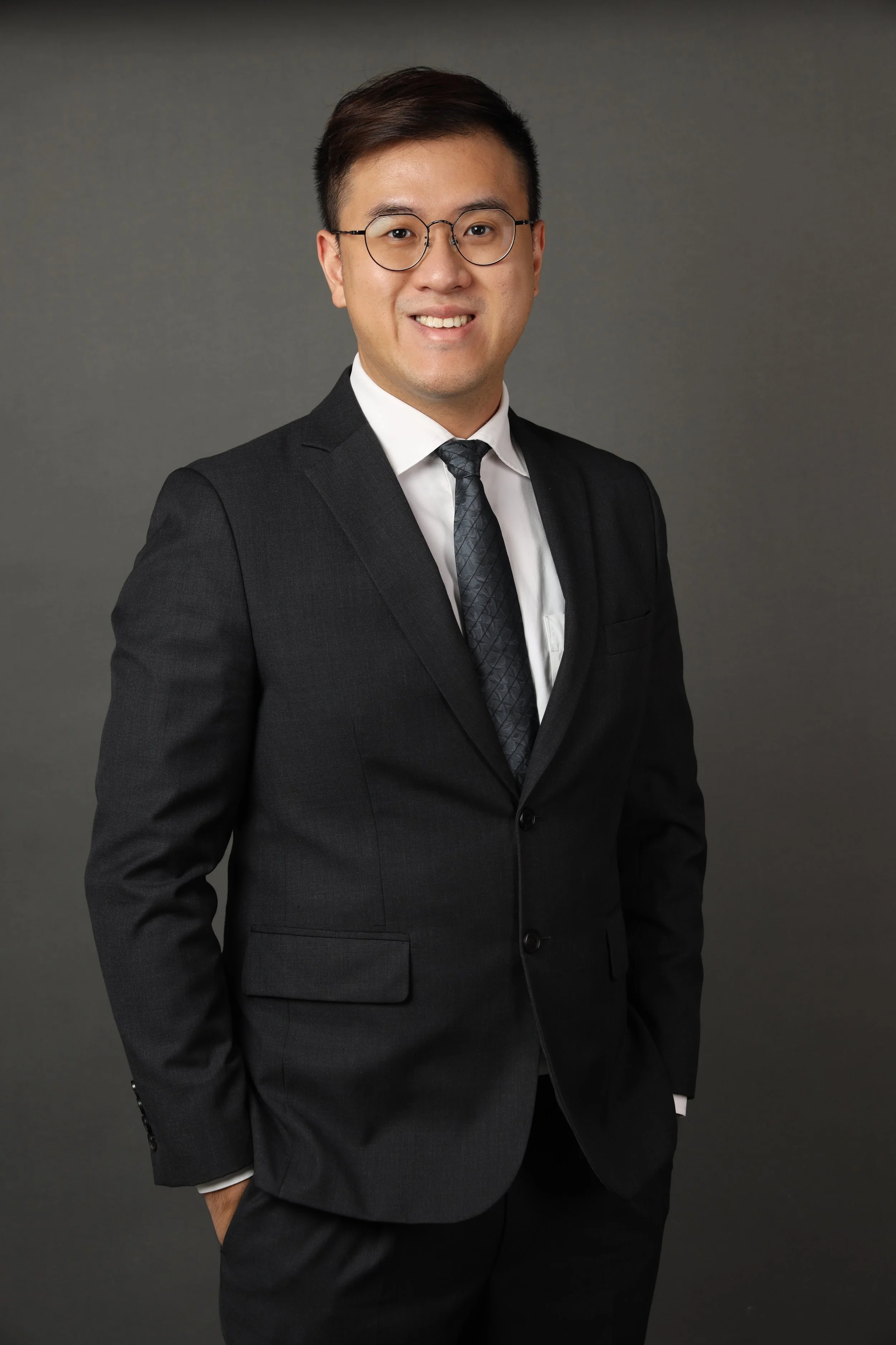 A professional man wearing glasses, a black suit, white shirt, and patterned black tie, standing against a gray background and smiling.
