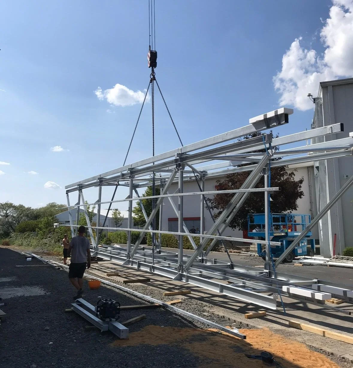 Construction workers install a large metal framework structure outdoors on a sunny day, with a blue sky and scattered clouds in the background.