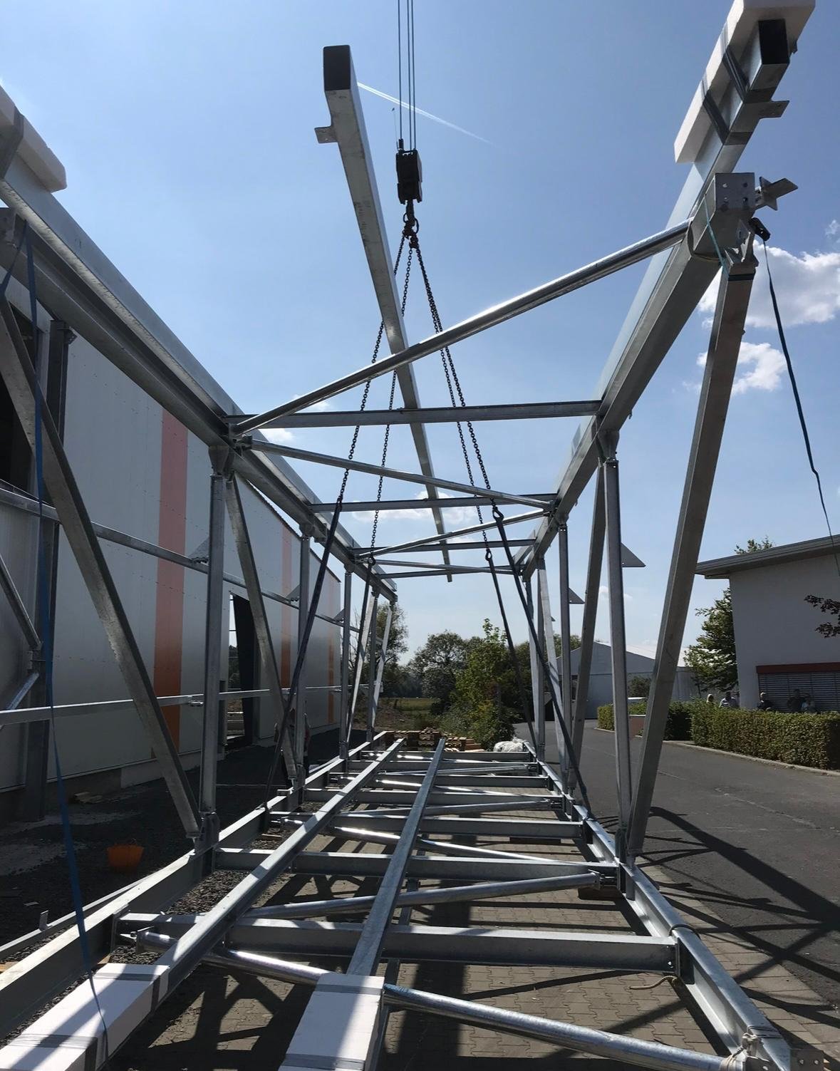 Metal framework structure, possibly for a satellite dish or large antenna, partially assembled outdoors on a paved area with a building and trees in the background.