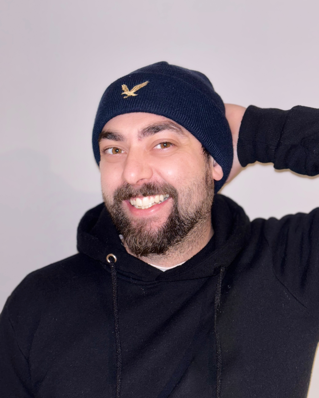 Ein lachender Mann trägt eine schwarze Kapuzenjacke und eine dunkle Mütze mit einem goldenen Vogel-Emblem, die er freundlich anlächelnd hat.