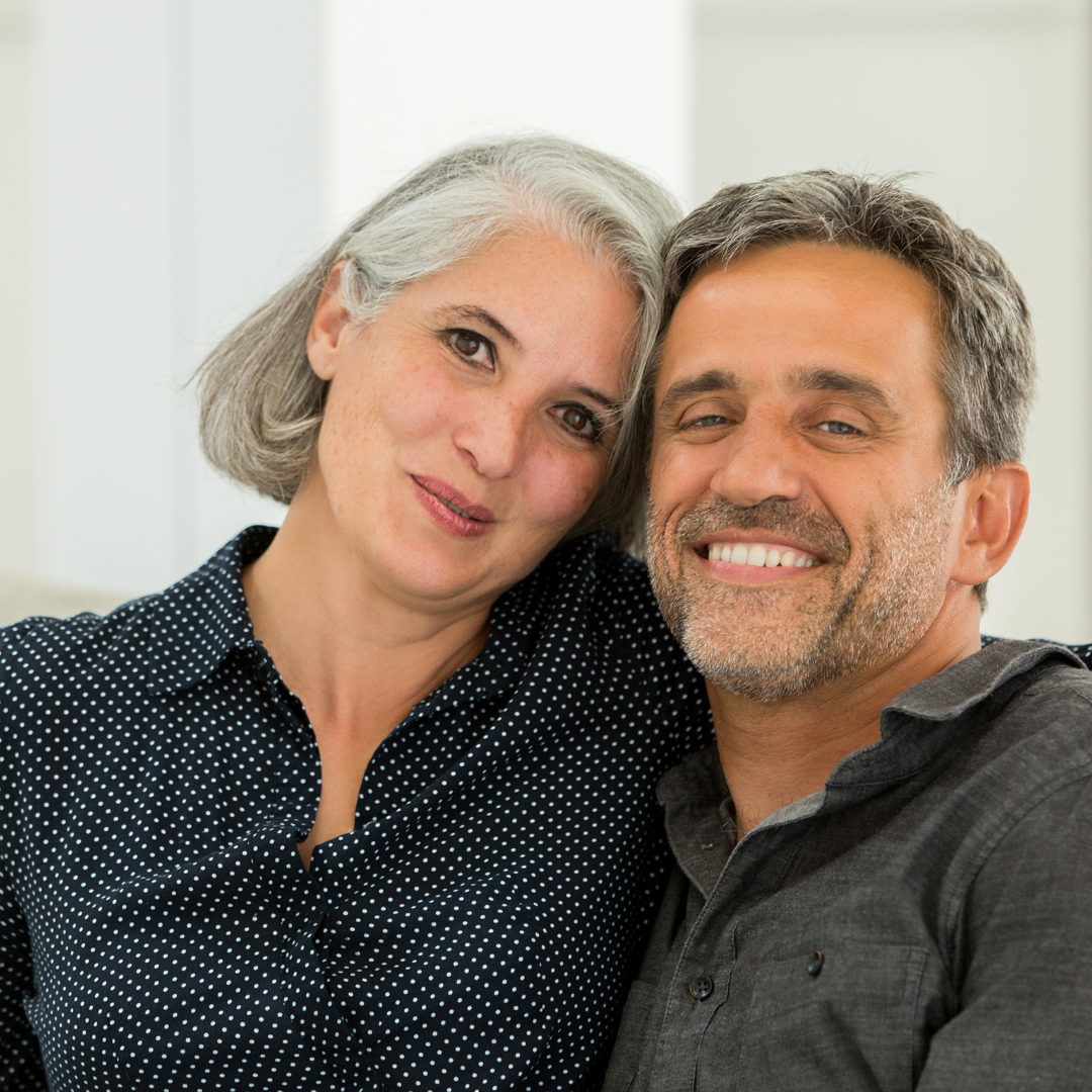 A smiling older couple sitting close together, the woman with gray hair and the man with dark hair and a beard, both casually dressed, in an indoor setting.