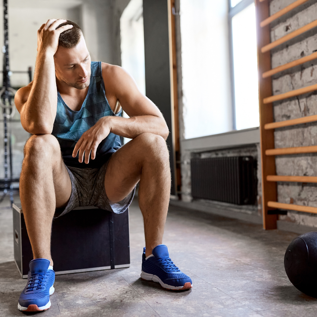 A tired man sitting on a workout box in a gym, resting after exercising, wearing a blue tank top, gray shorts, and blue athletic shoes.