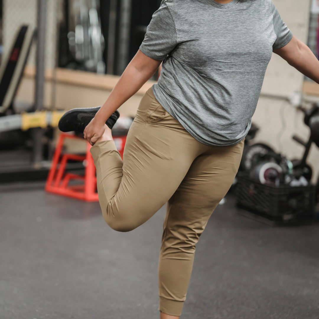 Person stretching in a gym, wearing a gray t-shirt and tan pants, holding one leg behind them for a stretch.