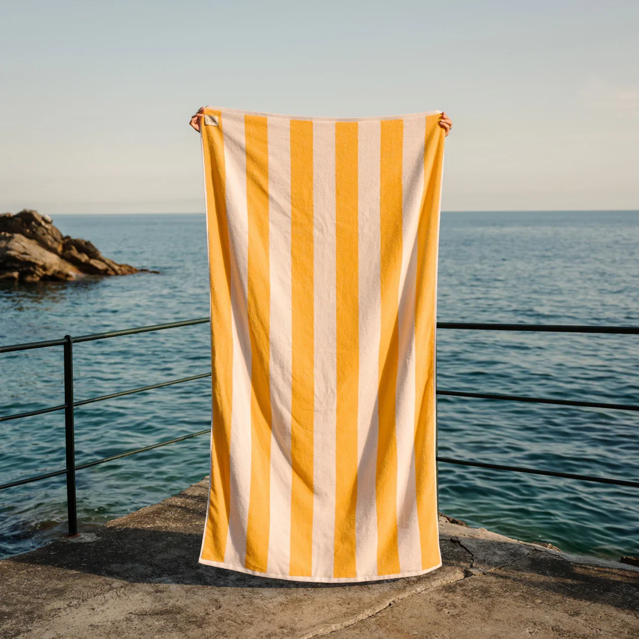 A person holding a yellow and white striped towel on a concrete pier by the ocean with rocks and a clear sky in the background.