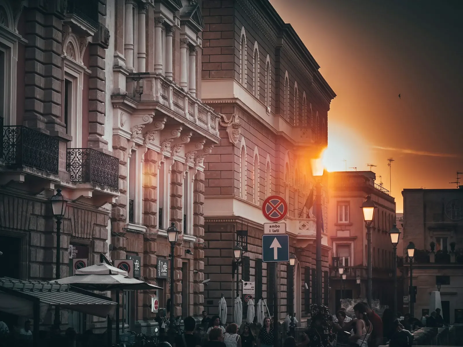Street scene at sunset on Piazza Sant'Oronzo with historic buildings, street lamps, and people sitting at outdoor cafes.